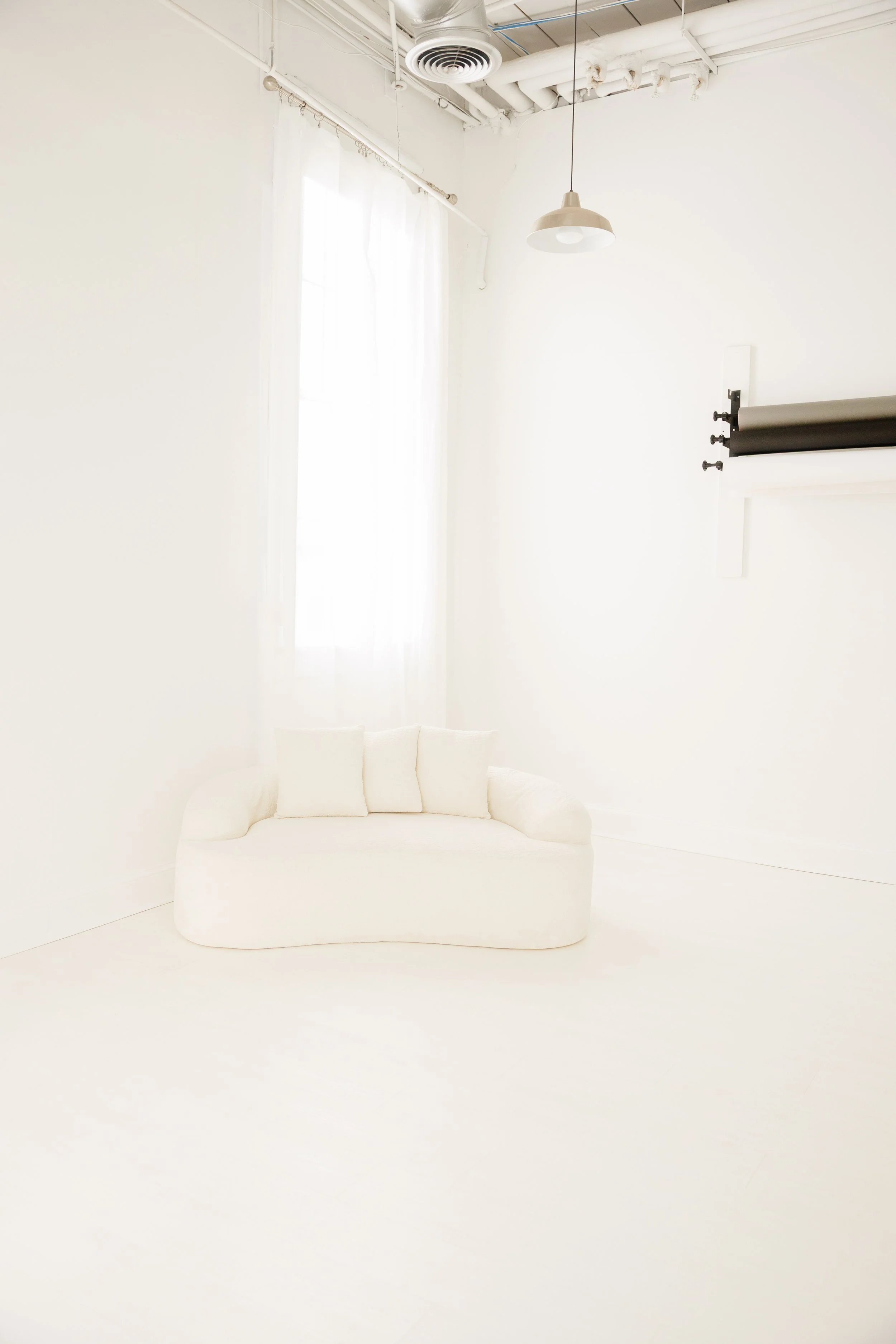 Bright white room with a small white sofa, sheer white curtains, and a photo studio backdrop holder on the wall.