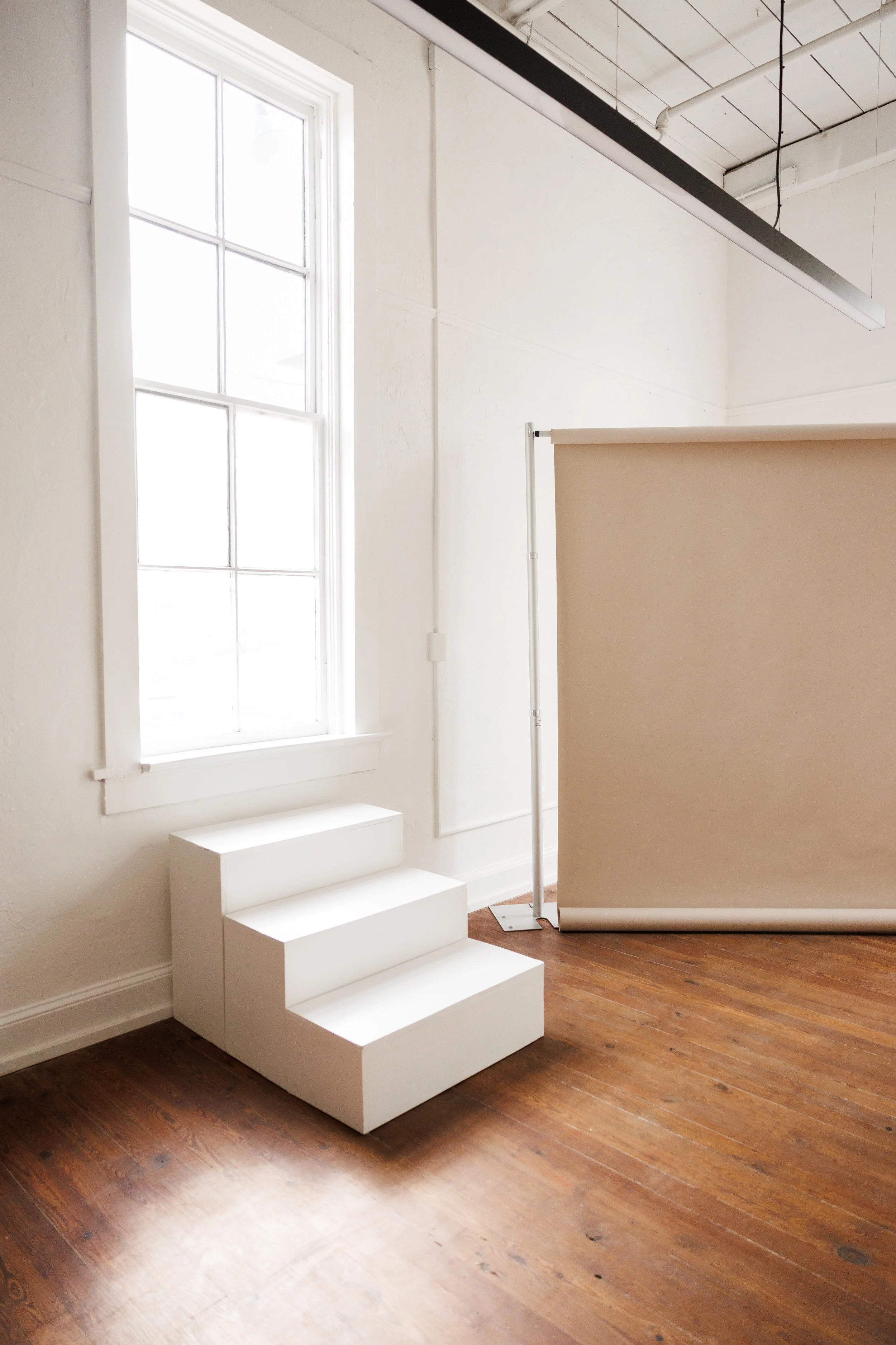 White interior room with wooden floor, large window, small white staircase, and a beige backdrop with a metal stand.