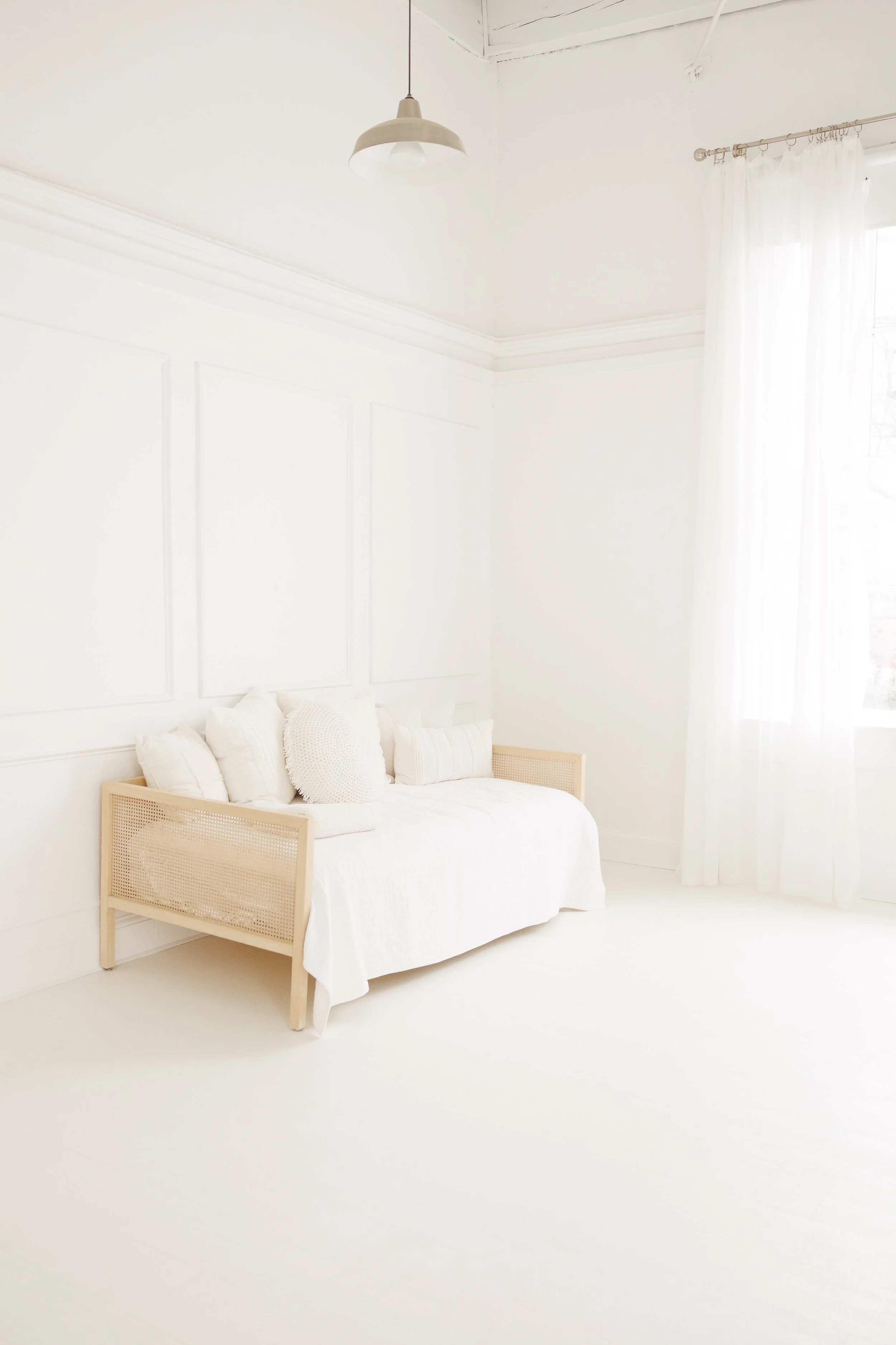 Bright, minimalist room with a white sofa adorned with white pillows, white curtains, a window allowing natural light, and a ceiling light fixture.