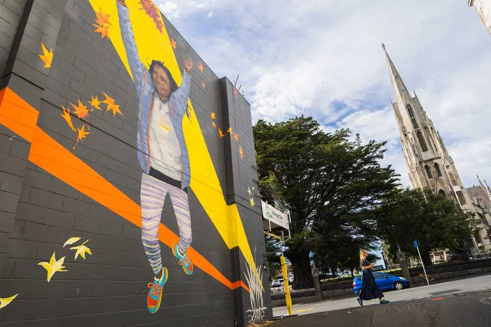 A colorful mural of a jumping child on an urban brick wall with trees, a cathedral, and a person walking nearby.