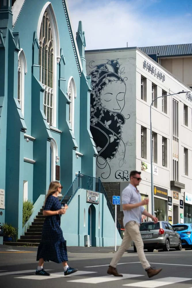 People walking across a crosswalk in front of colorful buildings, including a blue church with arched windows and a mural of a woman's face on the adjacent wall.