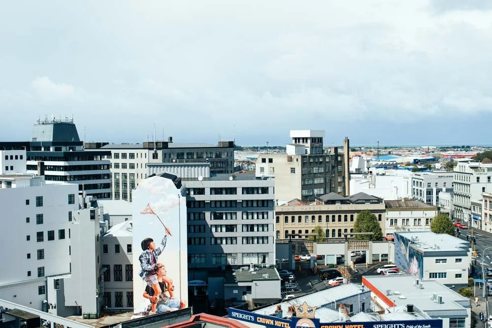 Cityscape with various buildings, a mural of children flying a kite on a building, and a cloudy sky.