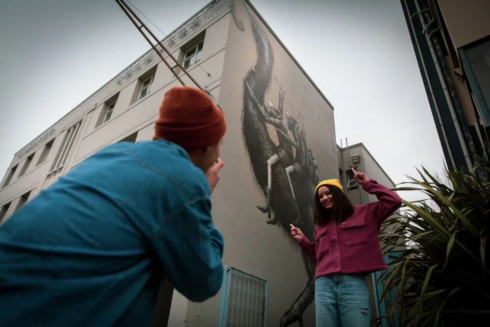 Two women are near a large building with a mural of a person climbing on it. One woman, wearing a yellow beanie and a maroon jacket, is pointing upward while smiling. The other woman, wearing a red beanie and blue jacket, is looking at her and appears to be taking a photo or looking up at the mural.