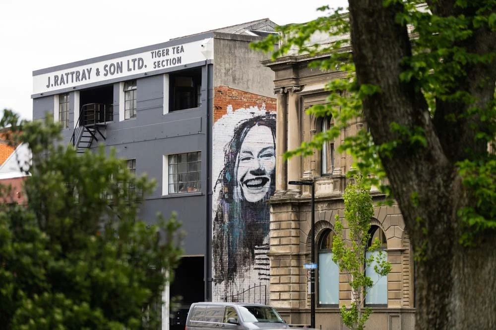 Large mural of a woman's face laughing on the side of a building, with trees and a parked van in the foreground.