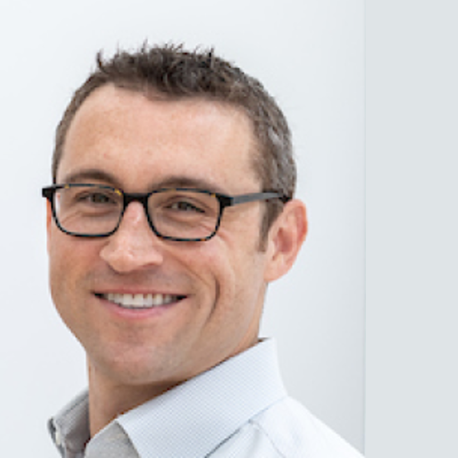 Portrait of a man with short brown hair, glasses, and a light blue collared shirt smiling in front of a white background.