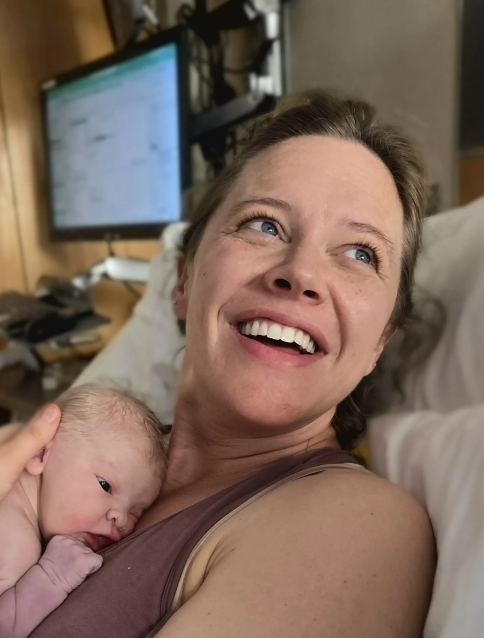 A woman lying in bed with a newborn baby resting on her chest, both smiling; a TV screen and wooden panel wall in the background.
