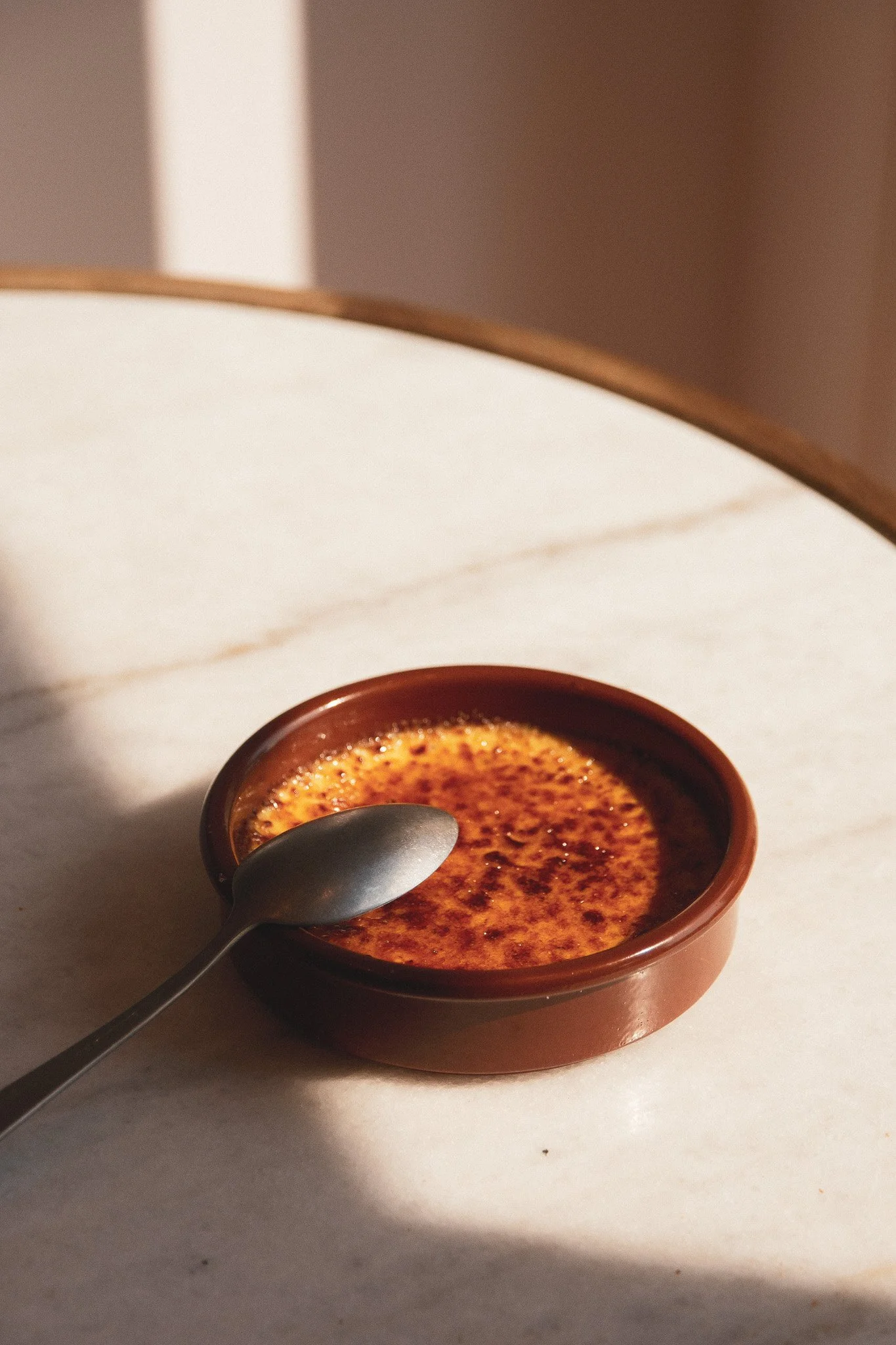 Crème brûlée dans un ramequin en terre cuite avec une cuillère à côté, placé sur une surface blanche, avec un fond flou.