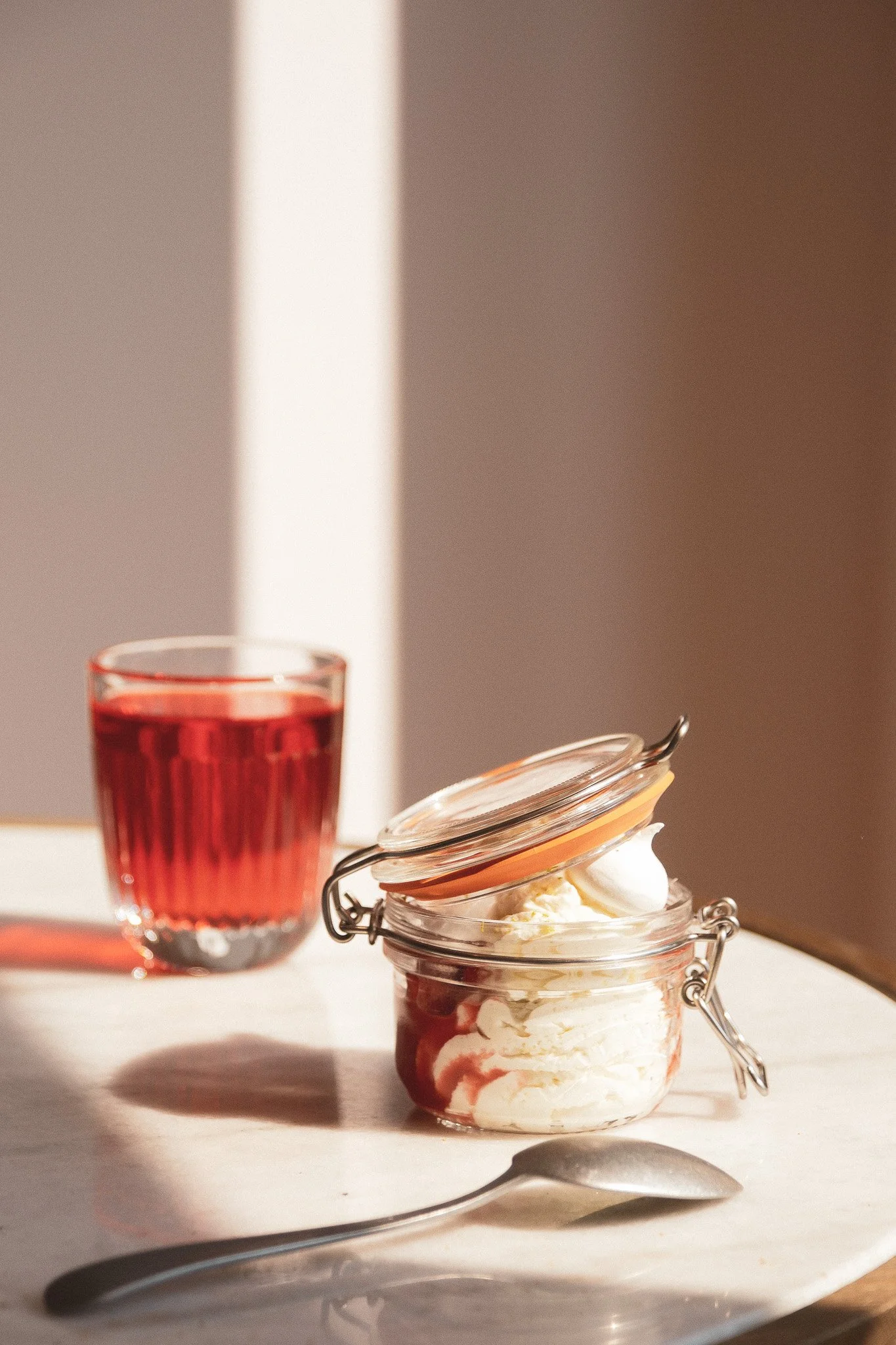 Un pot en verre contenant une coupe de glace à la vanille avec des morceaux de fruits rouges et une sauce rouge, une cuillère en métal à côté, une bouteille en verre avec une boisson rouge, et une paume en pierre sur une surface blanche, sous un écla