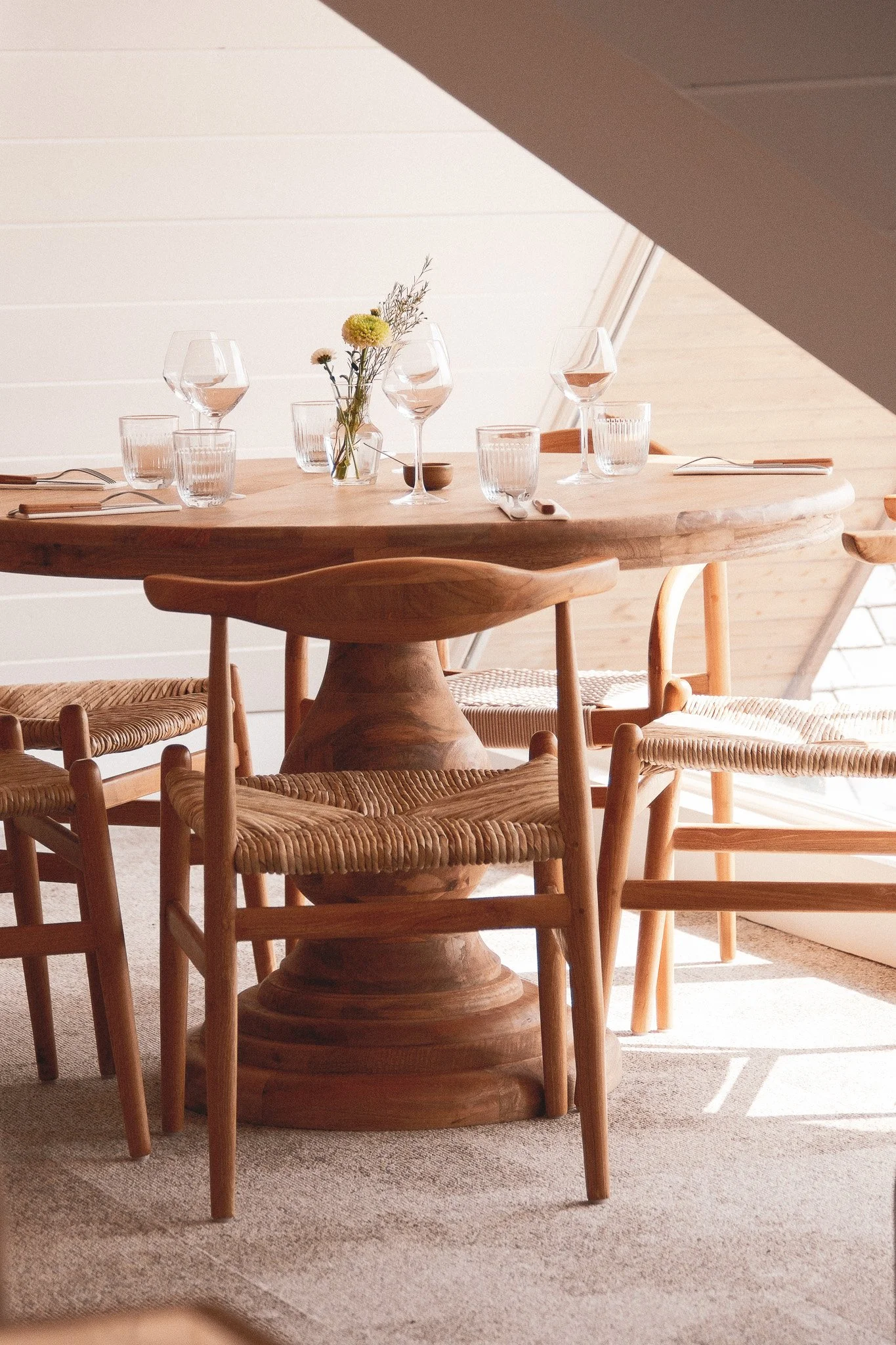 Table en bois avec des verres et une petite vase de fleurs à l'intérieur, entourée de chaises en bois avec assises en corde, dans une pièce bien éclairée avec un plafond en pente.
