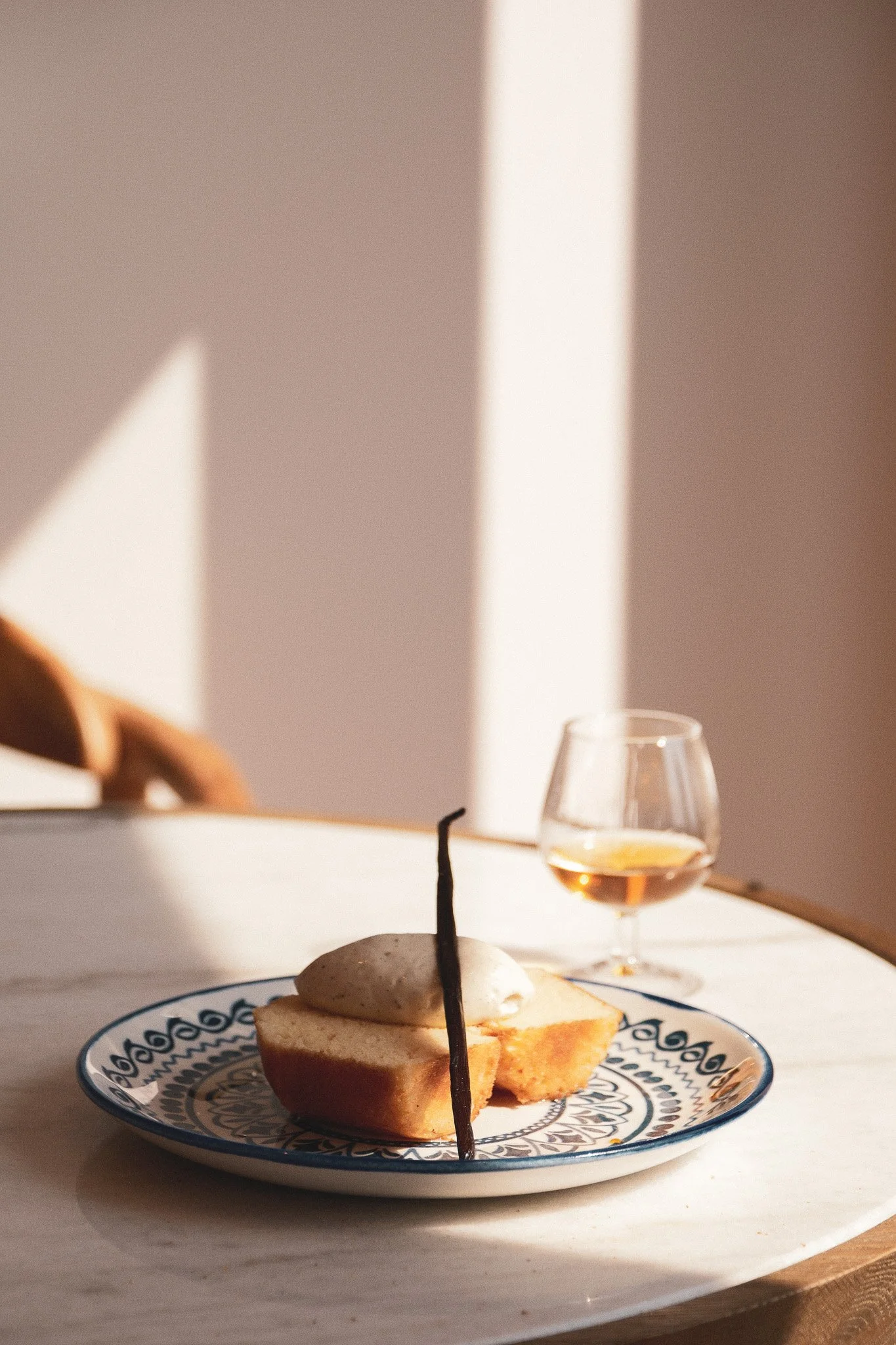 Une assiette avec un gâteau ou une pâtisserie, accompagnée d'un verre de vin ou de champagne, sur une table blanche avec une lumière naturelle ou artificielle douce.
