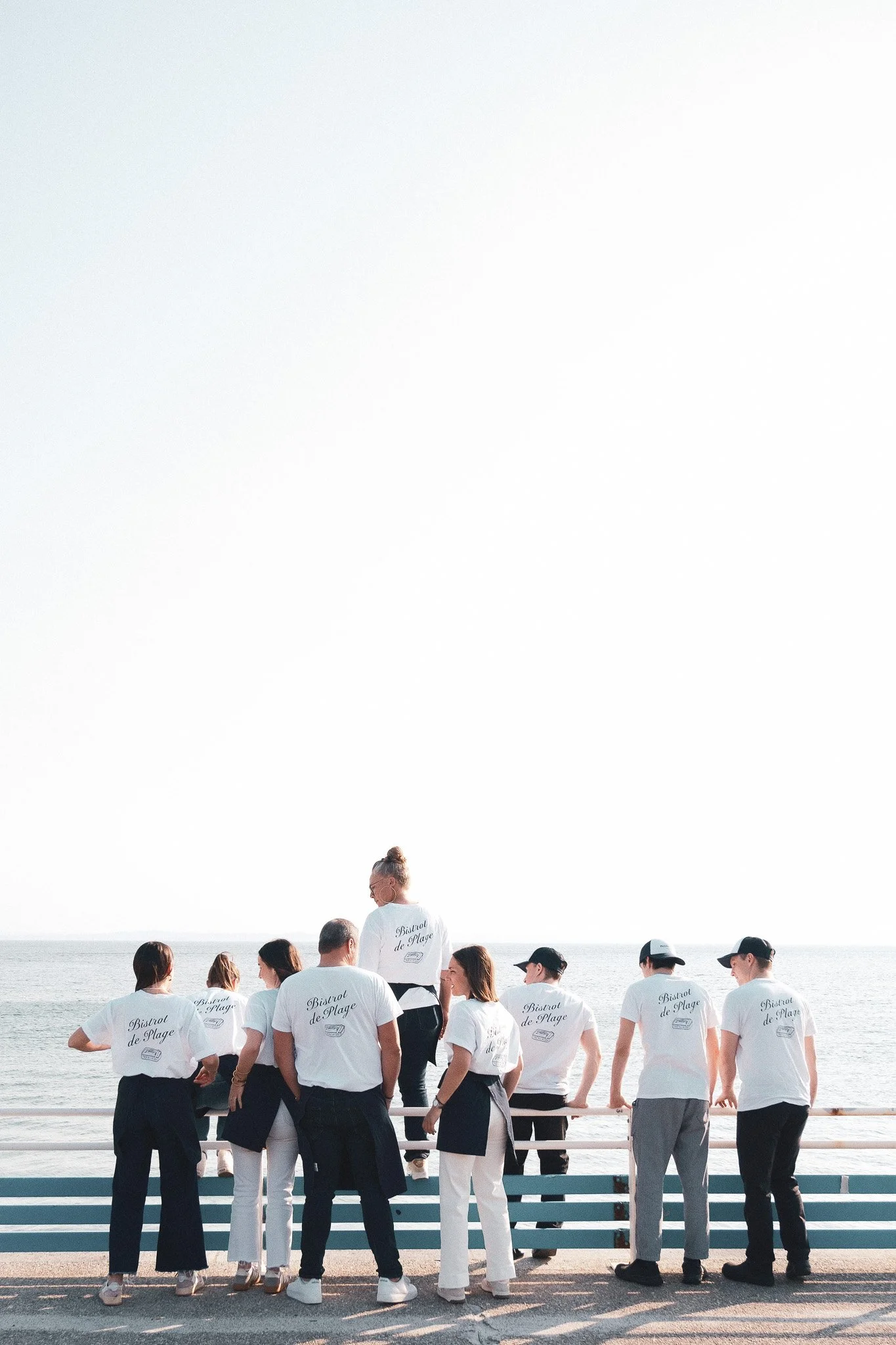Groupe de personnes portant des t-shirts blancs avec l'inscription 'Bistrot de Plage' sur la plage, face à la mer, sous un ciel clair.