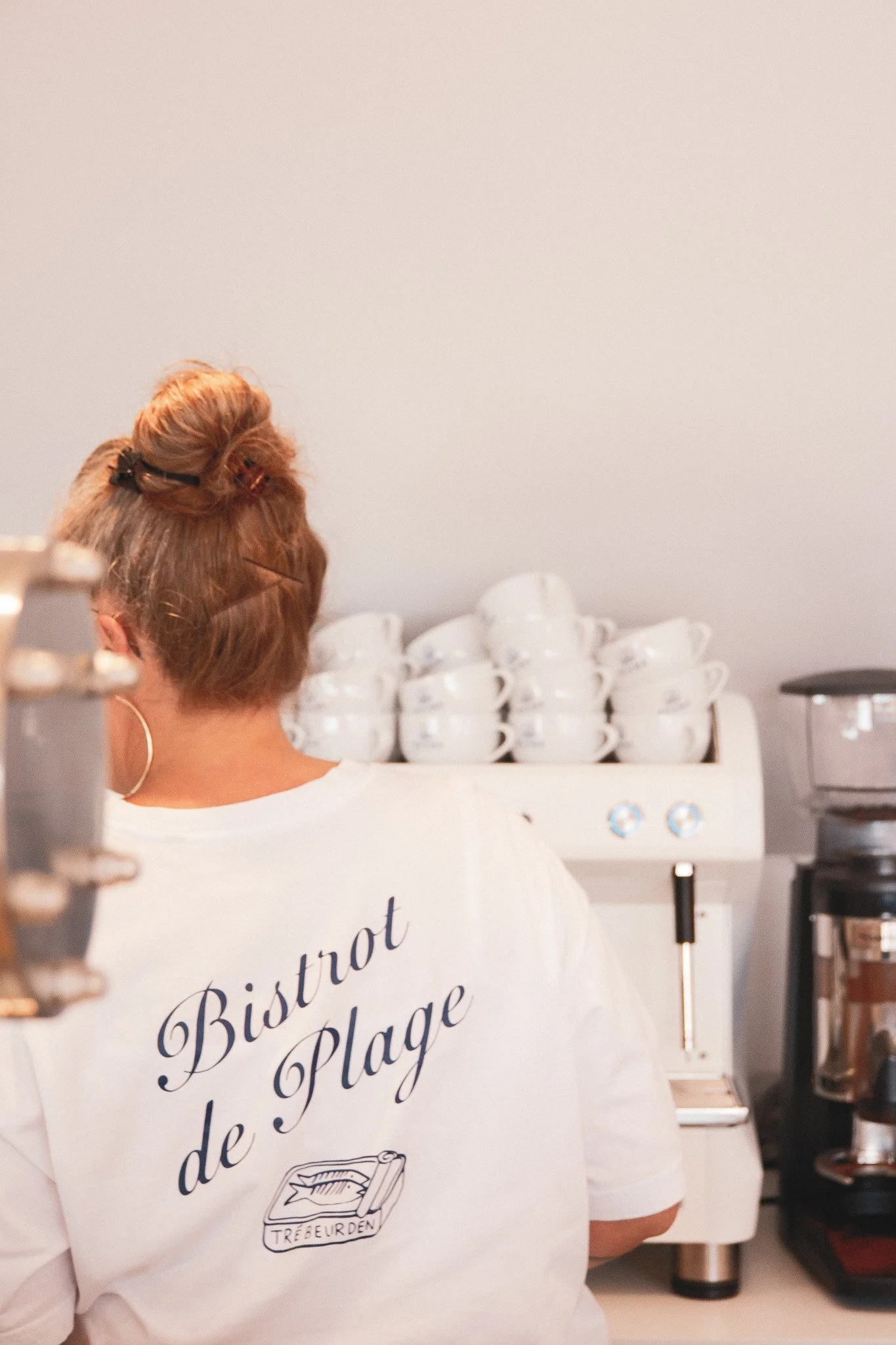 Une personne vue de dos dans une cuisine, portant un t-shirt blanc avec un logo et du texte en français. Sur le comptoir, des tasses blanches empilées, une machine à café, et un panneau indiquant 'Bistrot de la Plage'.