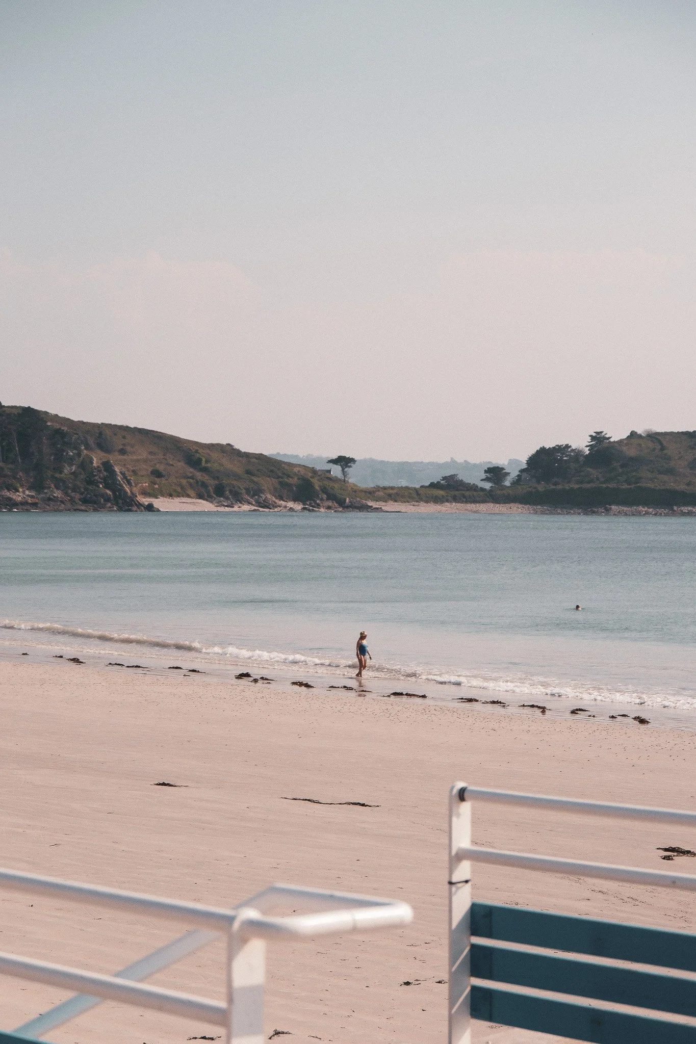 Plage de sable avec une personne marchant près de l'eau, quelques autres personnes dans la mer, et des collines verdoyantes à l'horizon