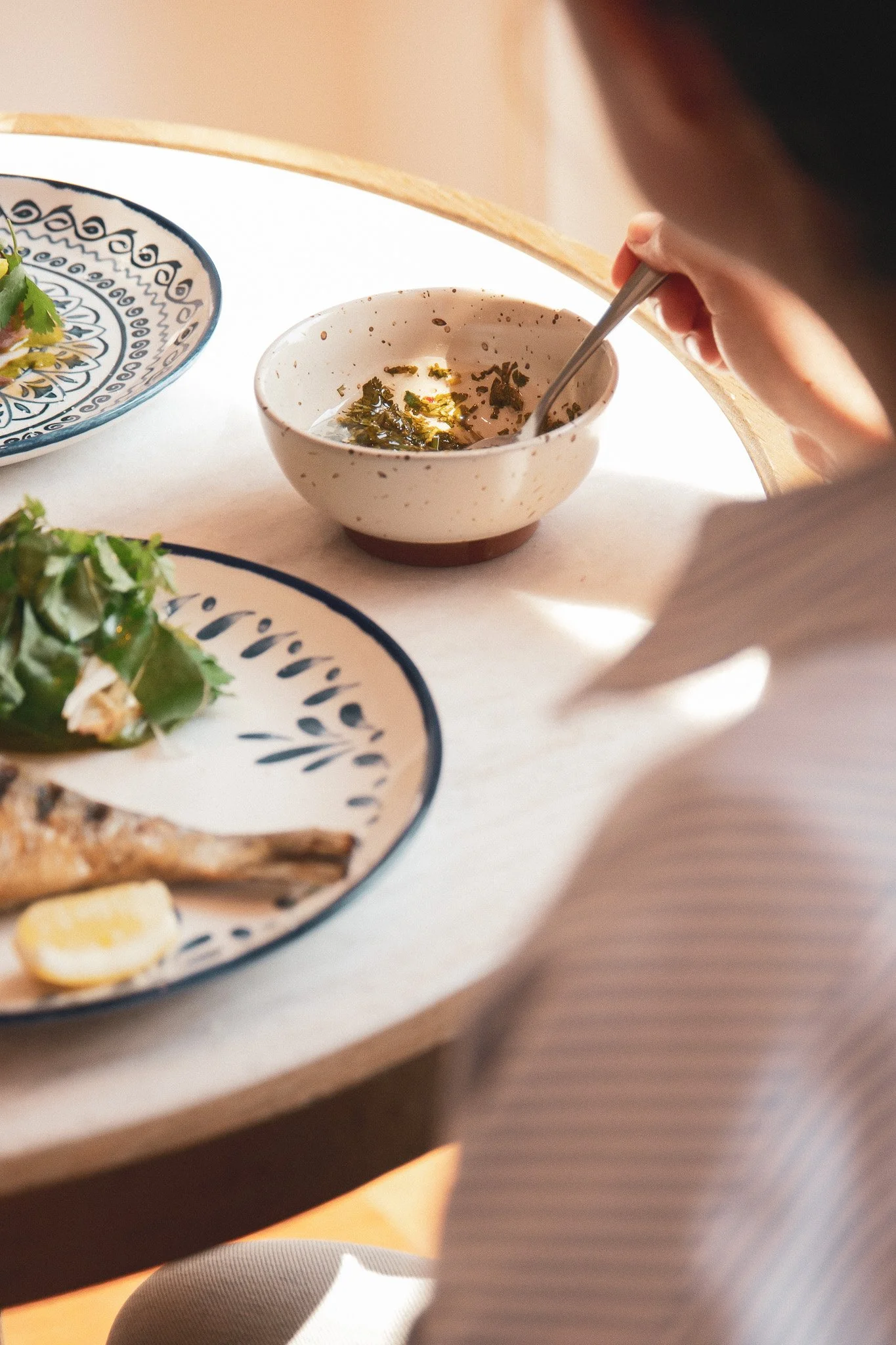 Une personne remuant une soupe dans un bol blanc avec des taches brunes, à côté de plats de nourriture sur une table en bois clair.