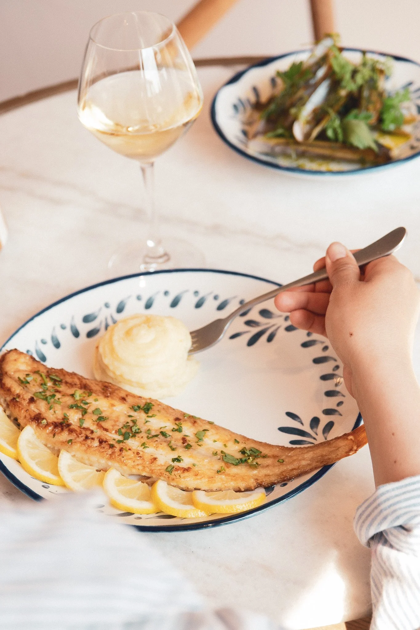 Assiette de poisson grillé garnie de tranches de citron, accompagnée de pommes de terre en purée, avec un verre de vin blanc et une salade verte en arrière-plan.