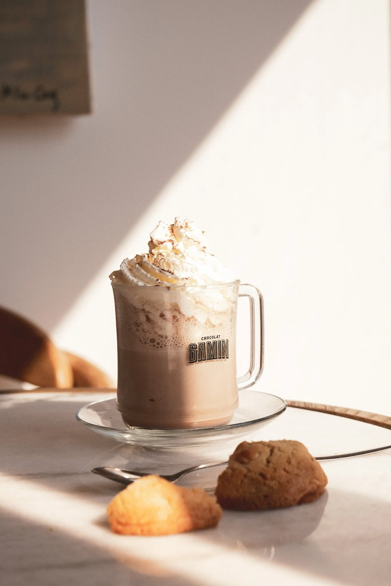 Une boisson chocolatée dans un verre en verre avec de la crème fouettée sur le dessus, une tablette de chocolat à l'intérieur du verre, une assiette avec deux biscuits et une cuillère, le tout sur une table blanche avec un fond clair.