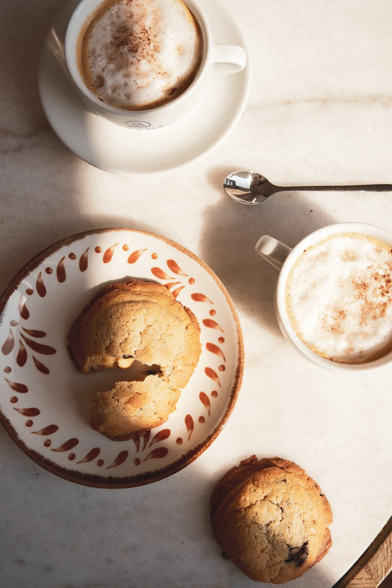 Une tasse de chocolat chaud ou de cappuccino avec de la mousse, une assiette avec deux cookies aux pépites de chocolat, et une cuillère en métal sur une surface en marbre clair.