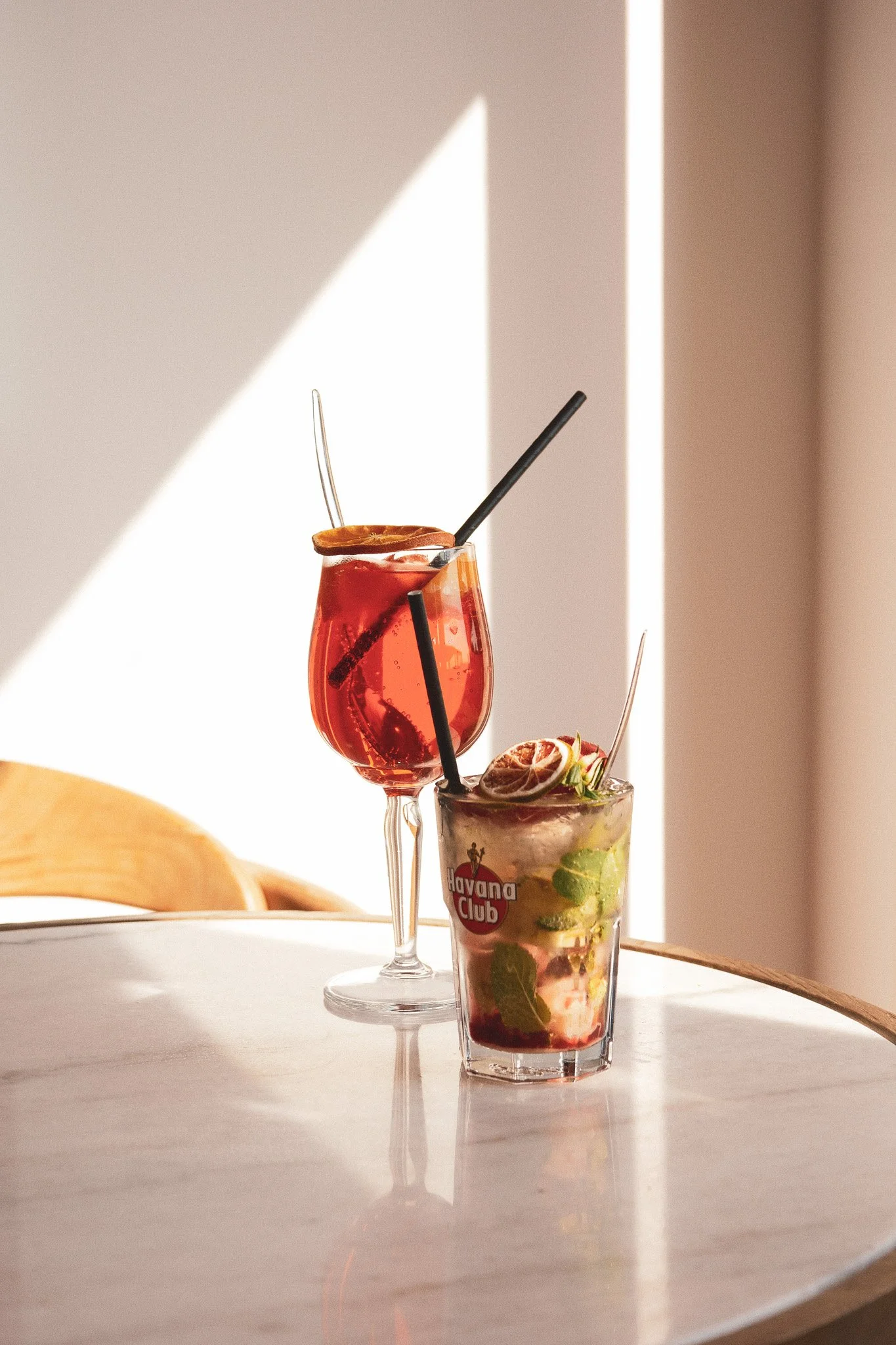Deux cocktails colorés sont placés sur une table ensoleillée, l'un avec une garniture d'orange, l'autre avec des tranches de fruits et des feuilles de menthe, dans un décor lumineux avec un mur clair en arrière-plan.
