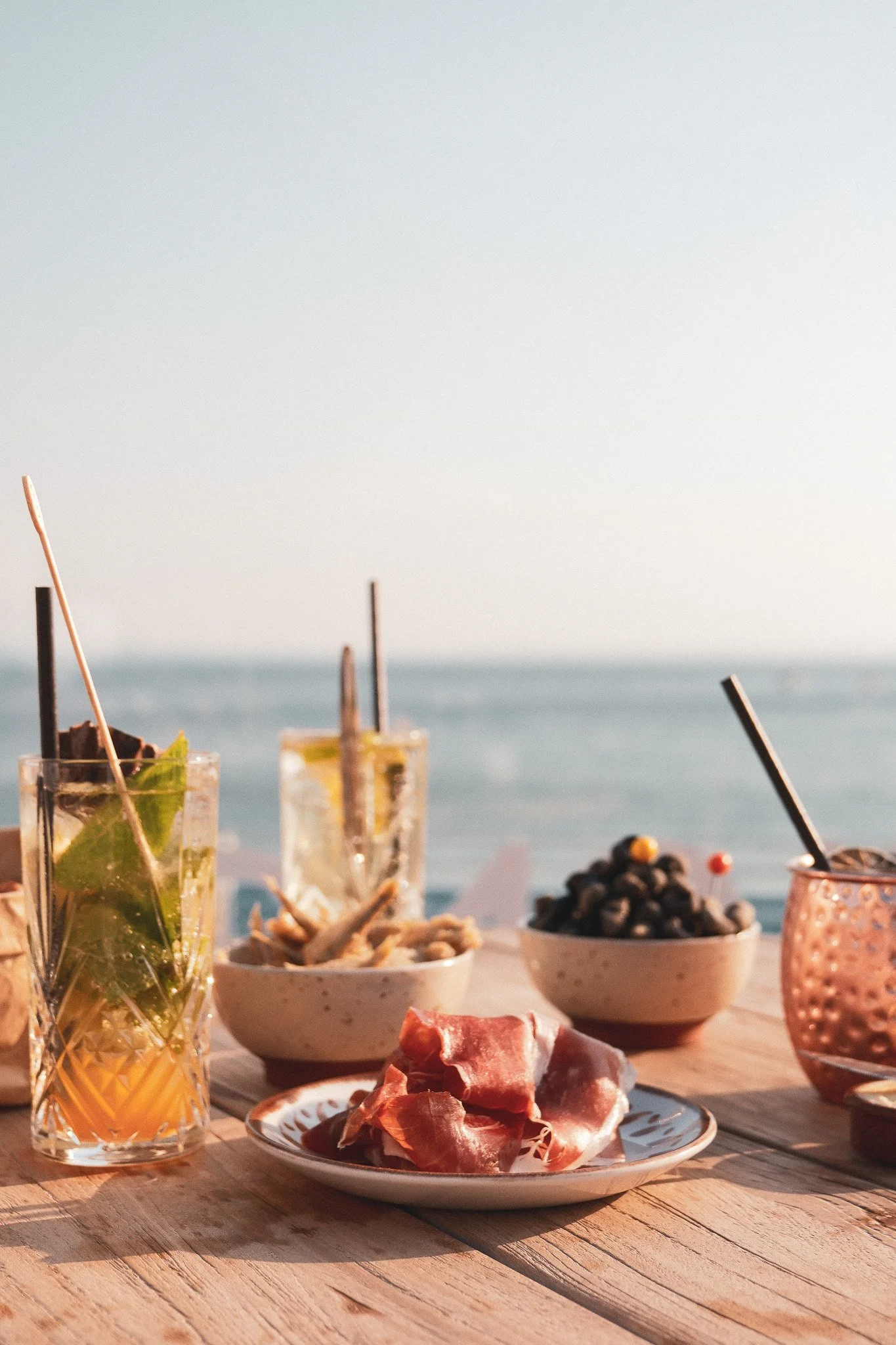 Assortiment de boissons et de snacks sur une table en bois face à la mer, avec un ciel clair.