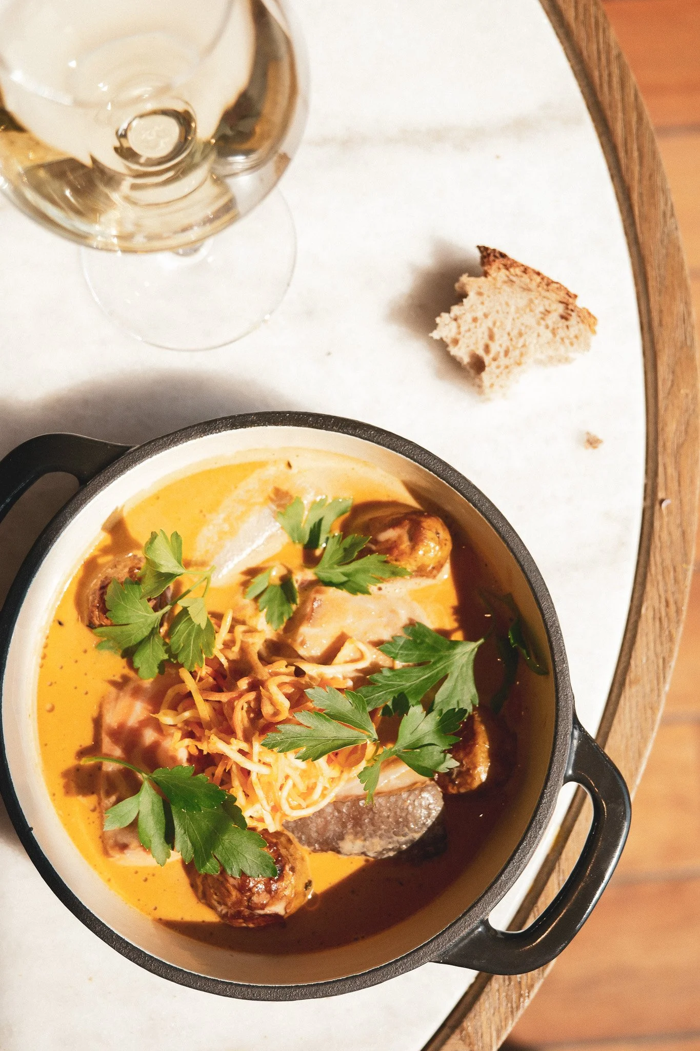 Soupe ou curry dans un plat noir avec des morceaux de viande, des herbes et un peu de fromage râpé, avec un verre de vin blanc en arrière-plan et un morceau de pain à côté.