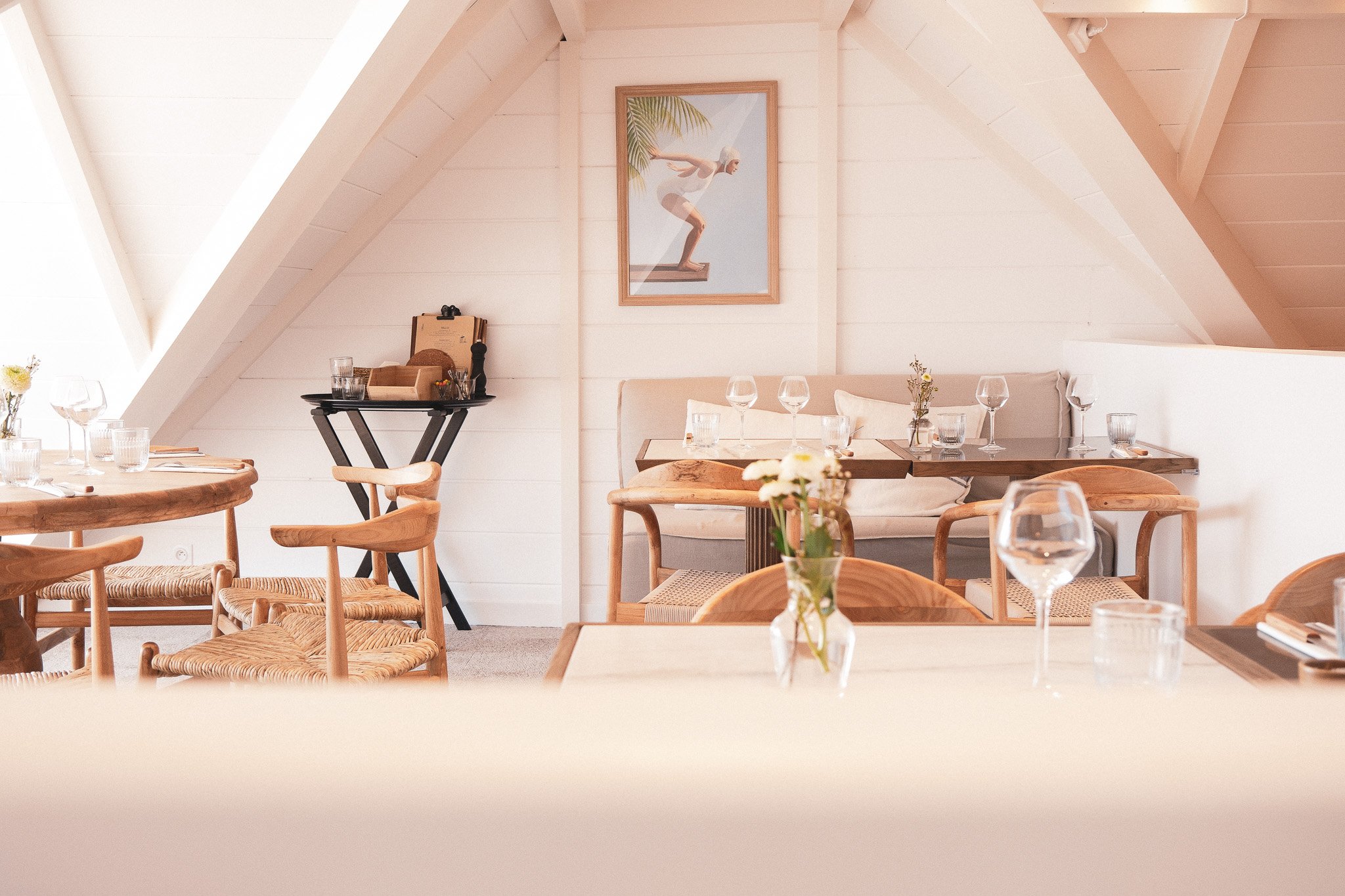 Salle à manger chaleureuse avec tables en bois, verres et petite décoration florale, peinture de nageuse en arrière-plan, ambiance lumineuse et accueillante.