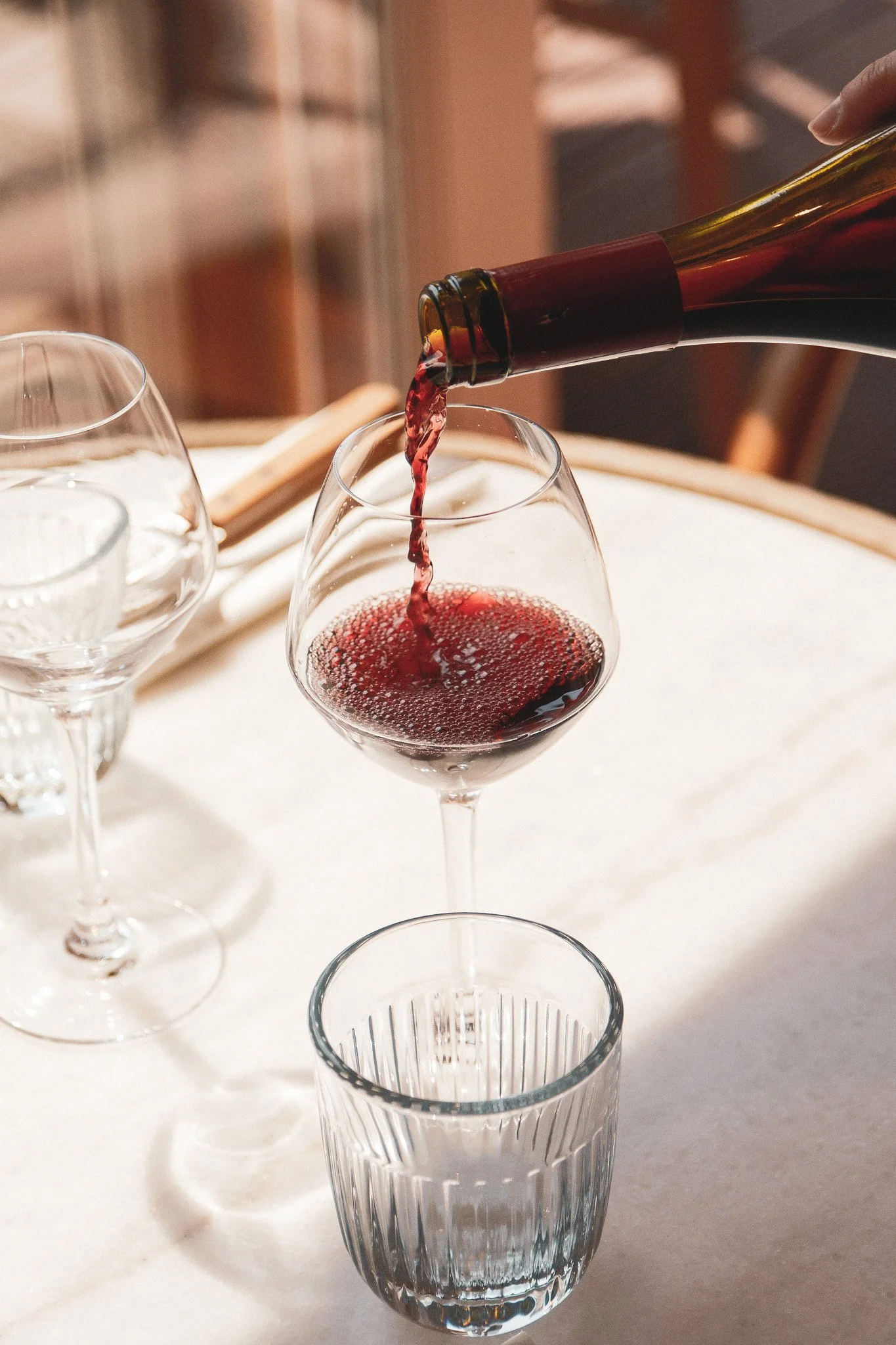Une main verse du vin rouge dans un verre à vin sur une table, avec plusieurs autres verres vides autour.