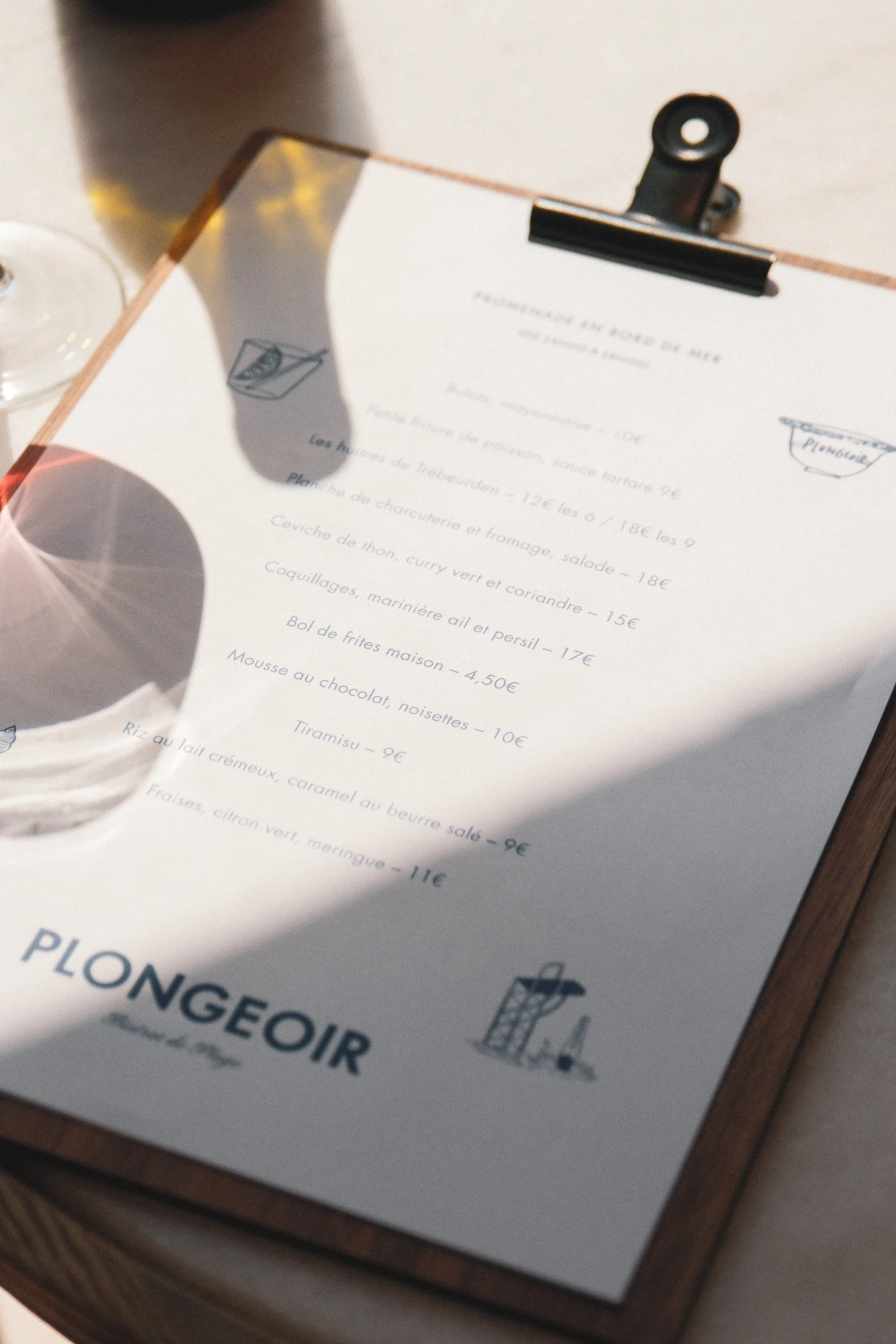 Une feuille de menu de restaurant avec des plats comme le poisson, le poulet, des desserts, et des illustrations de cuisines. Inclut une pince à papier pour maintenir la feuille. La feuille repose sur une surface en bois.