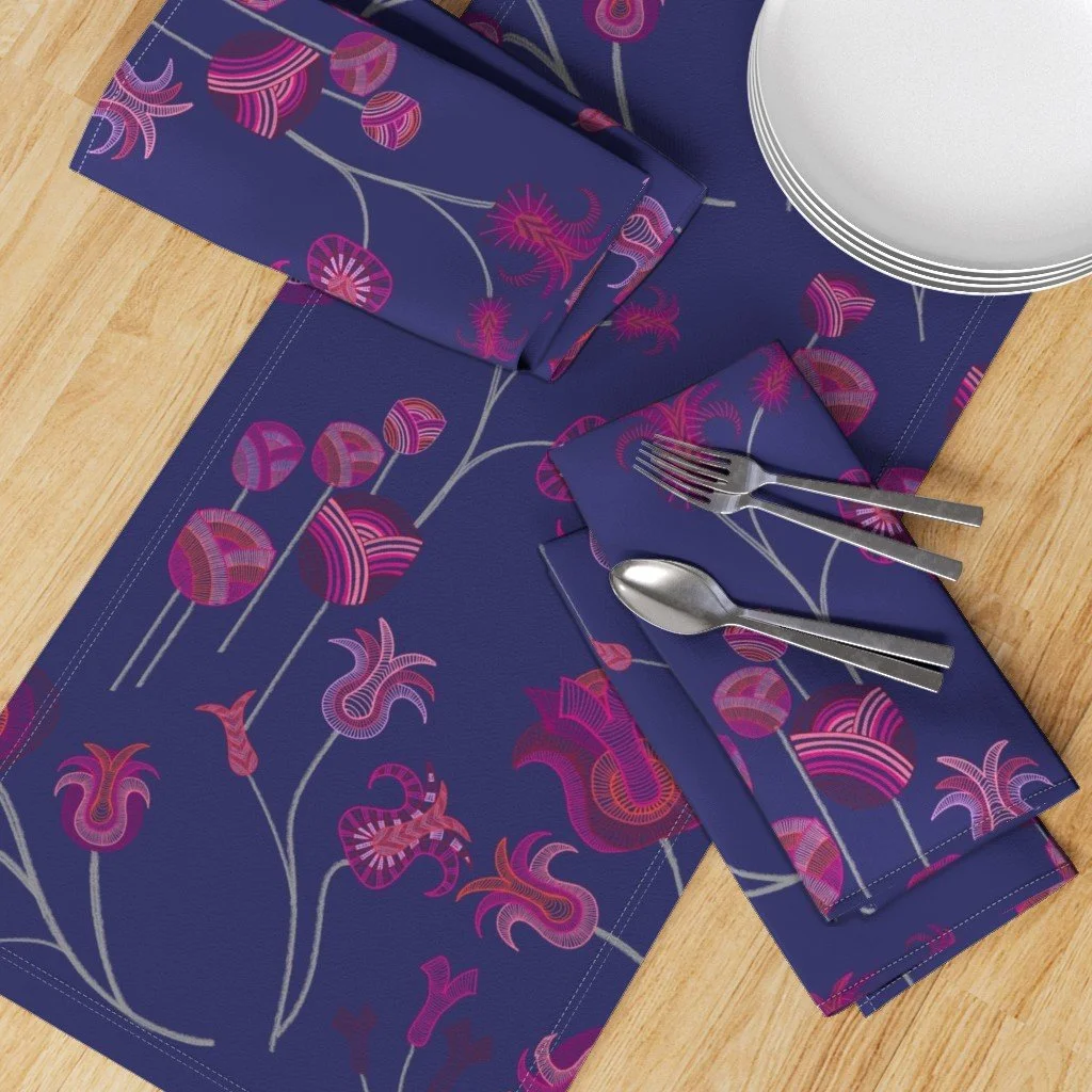 A table runner and napkins showing whimsical tulips that meander and sprout with detailed vibrancy against a deep blue ground sit atop a wood table with cutlery and a plate