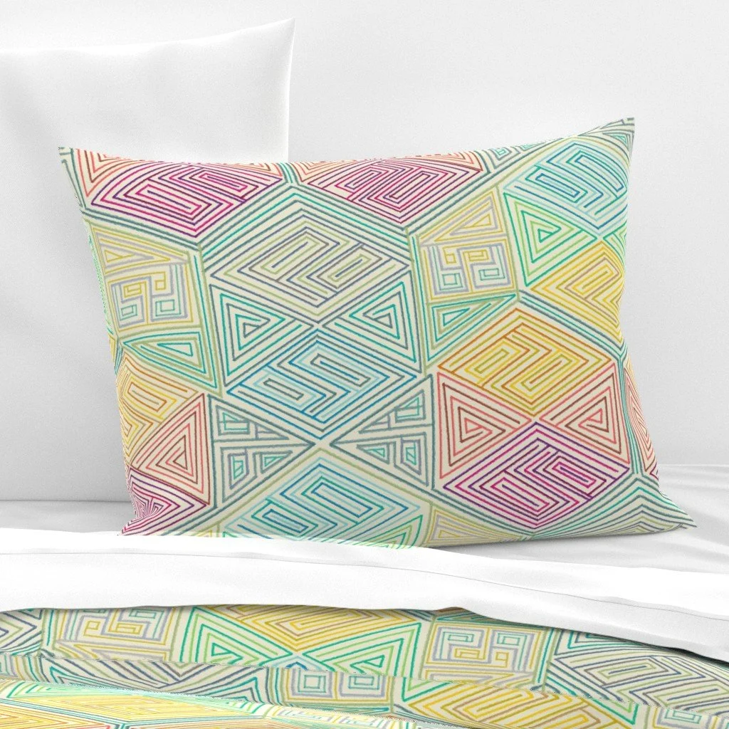 Rainbow hexagon maze pattern in hand-drawn line work covers a pillow sham propped against white bedding, with the same print echoed on the bed below.