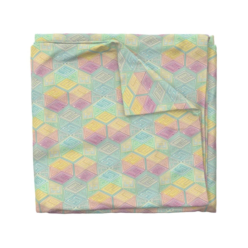 Rainbow hexagon maze pattern folds across a bolt of fabric, one corner turned back to reveal the print continuing beneath.