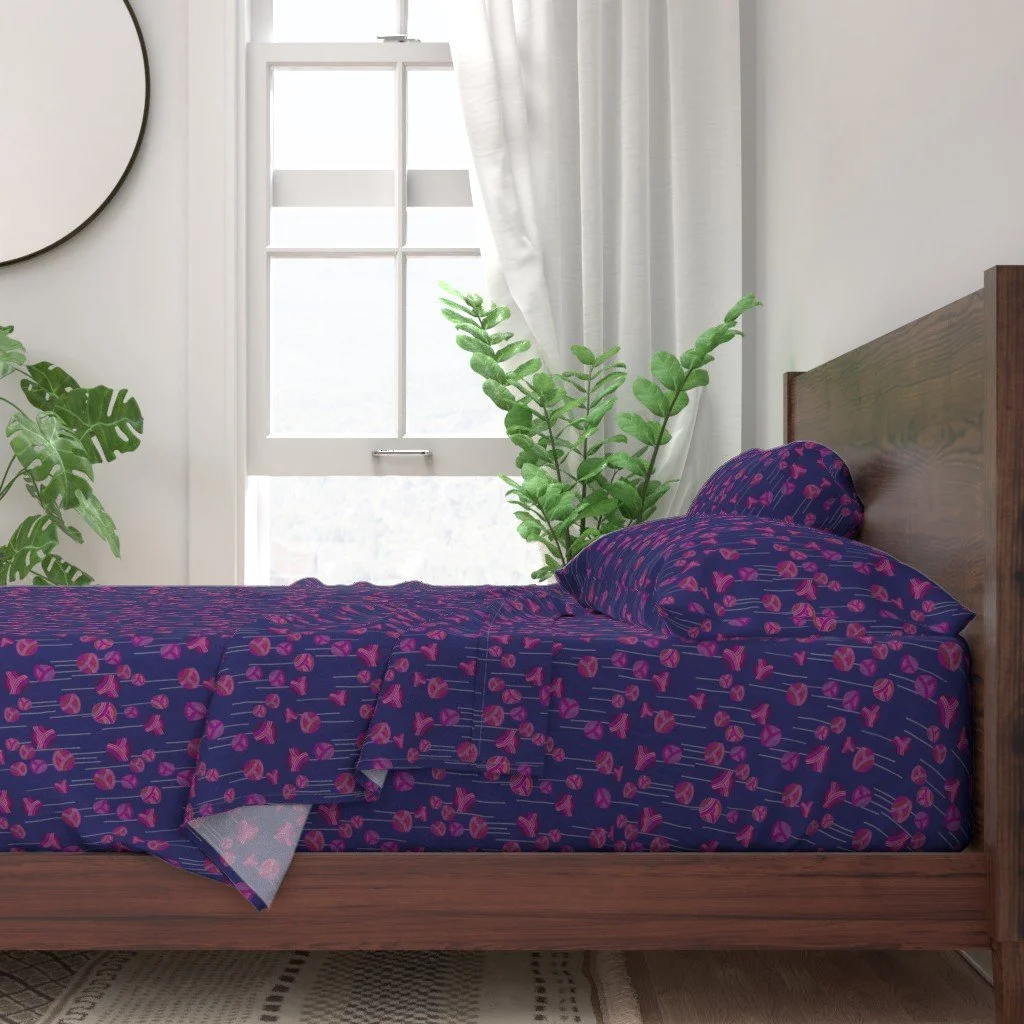 Sheets with a dancing floral ditsy pattern with abstract tulips and stems on a vibrant dark blue base sit upon a modern wooden bed in front of a white wall and bright window with plants in front of it