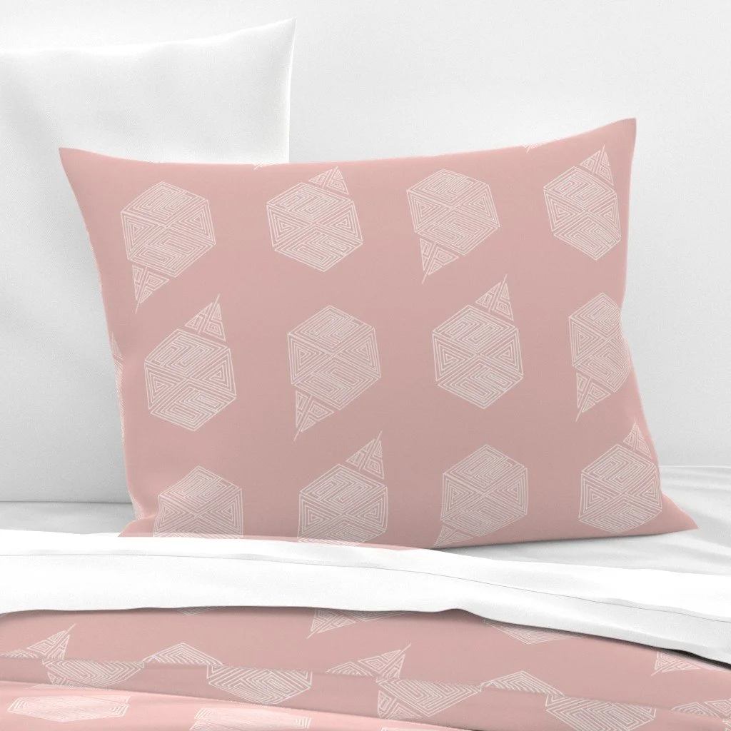 Blush pink geometric hexagon fabric shown on a bed pillow sitting in front of a pristine white wall and a white pillow, resting atop a bed with a matching top sheet, folded back.