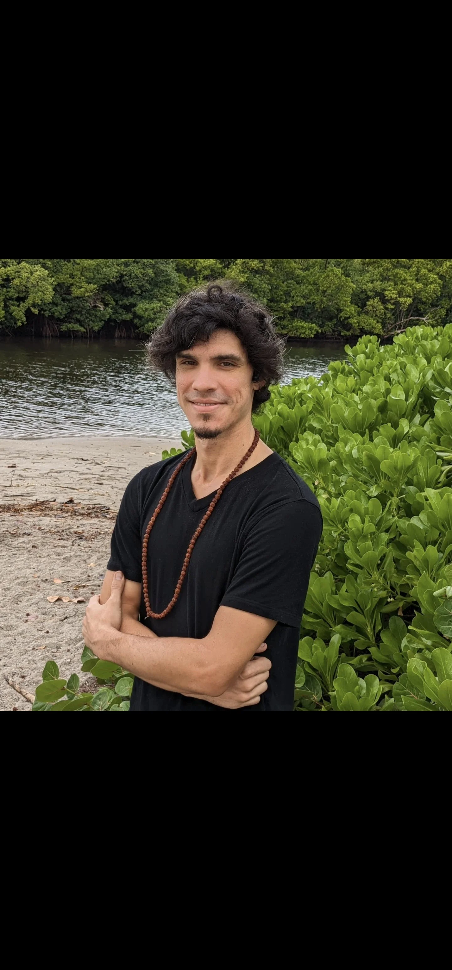 A man with curly dark hair, wearing a black t-shirt and a beaded necklace, stands on a beach with a river and lush green trees in the background.