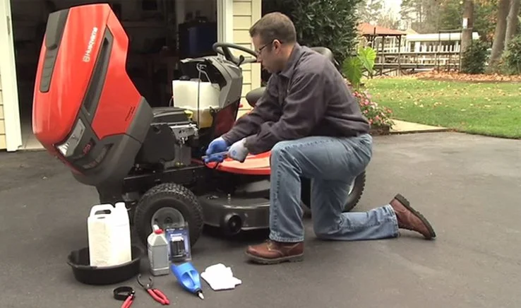 A man kneeling on the driveway, inspecting or repairing a red ride-on lawn mower, with various tools and supplies nearby.