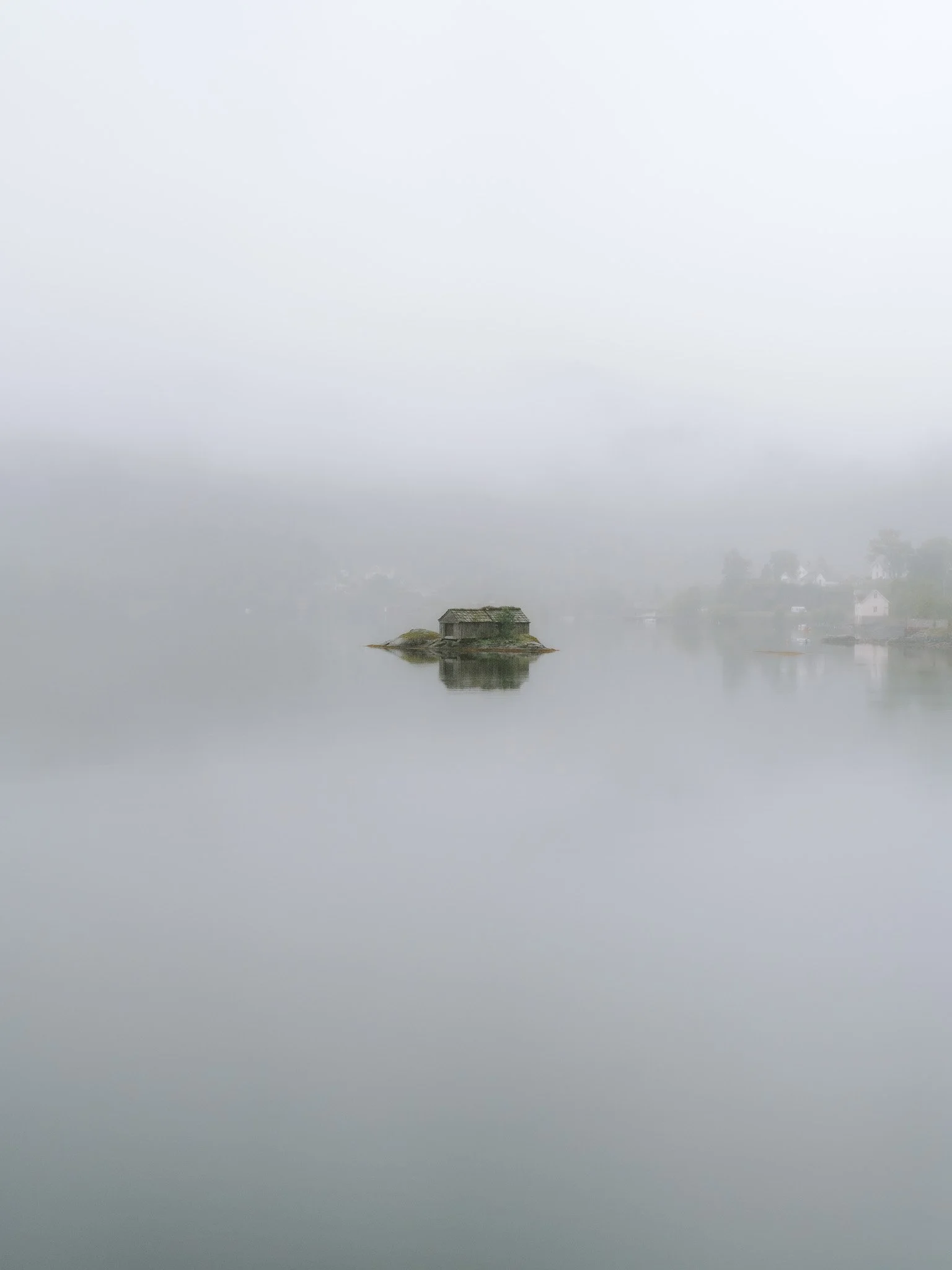 A small house on a tiny island in a foggy lake with calm water reflecting the house.
