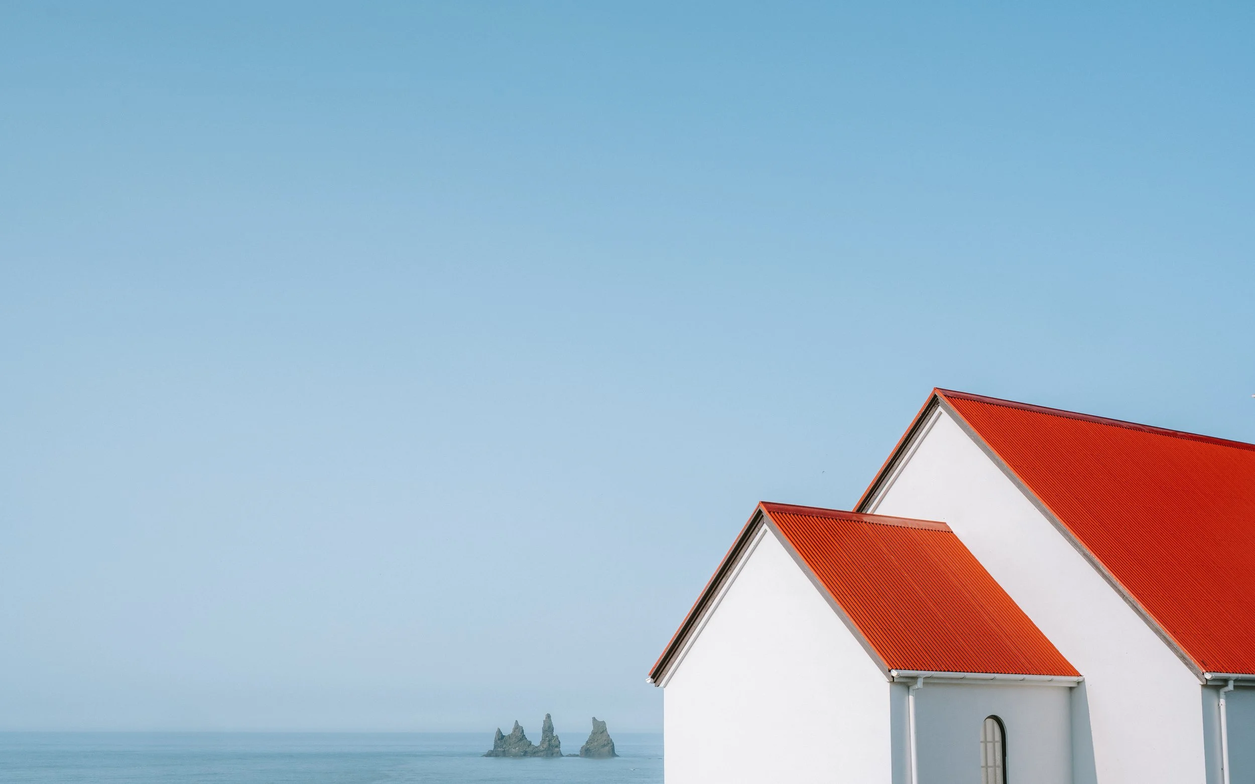White building with red gable roof against blue sky, ocean, and rock formations in the distance.