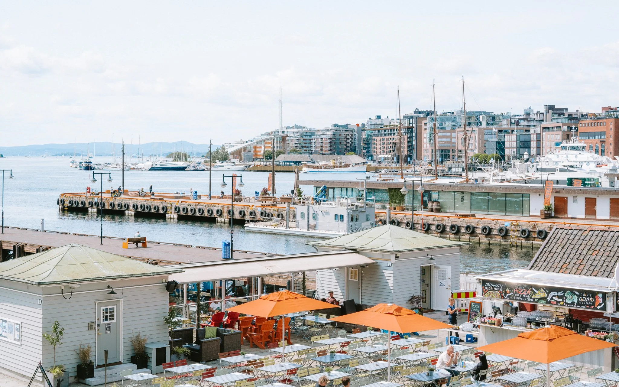 People dining outdoors with orange umbrellas near a waterfront with boats and city buildings in the background.