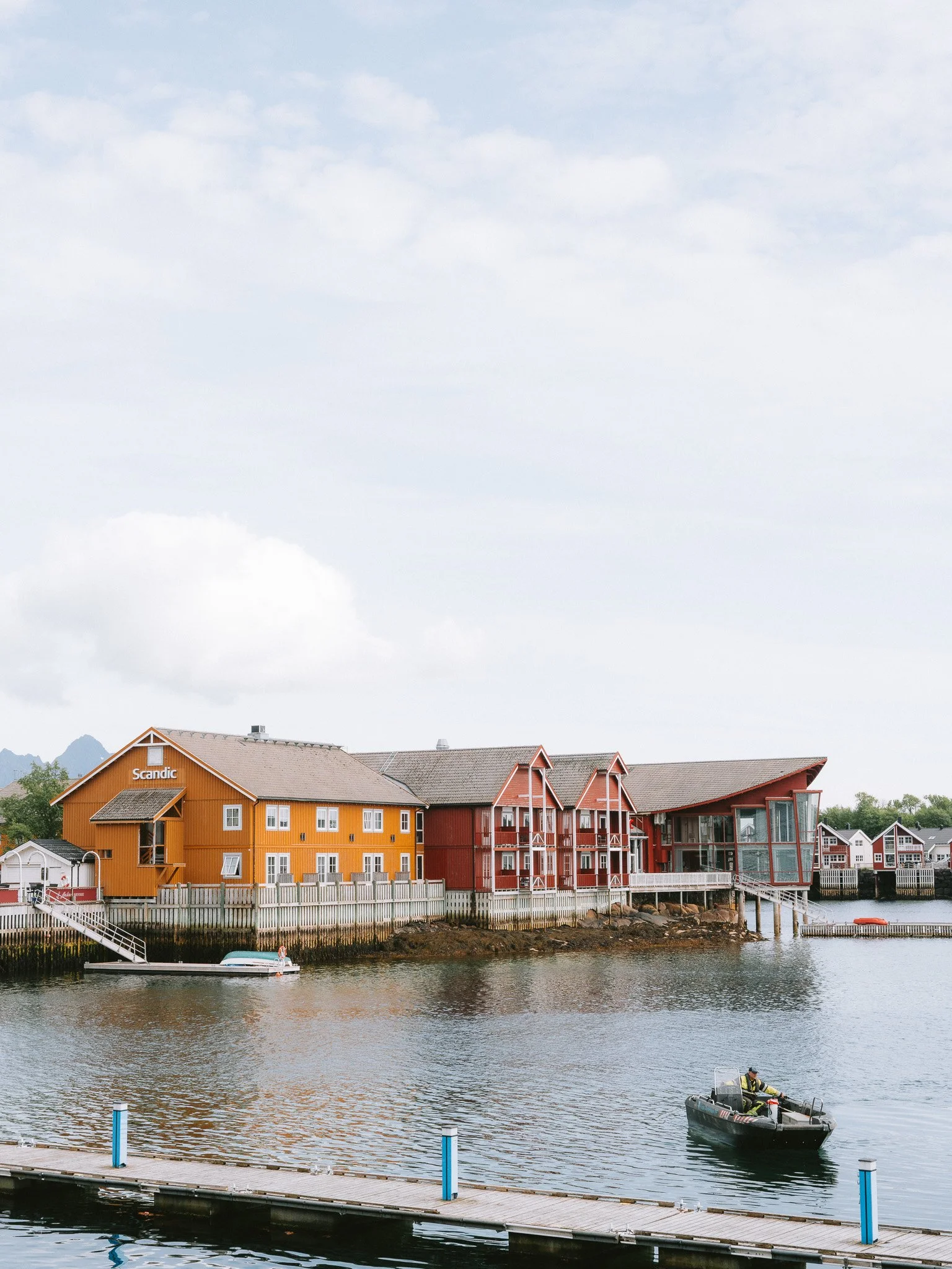 Colorful waterfront buildings, including orange and red structures, with a dock and a boat on the water, under a cloudy sky.