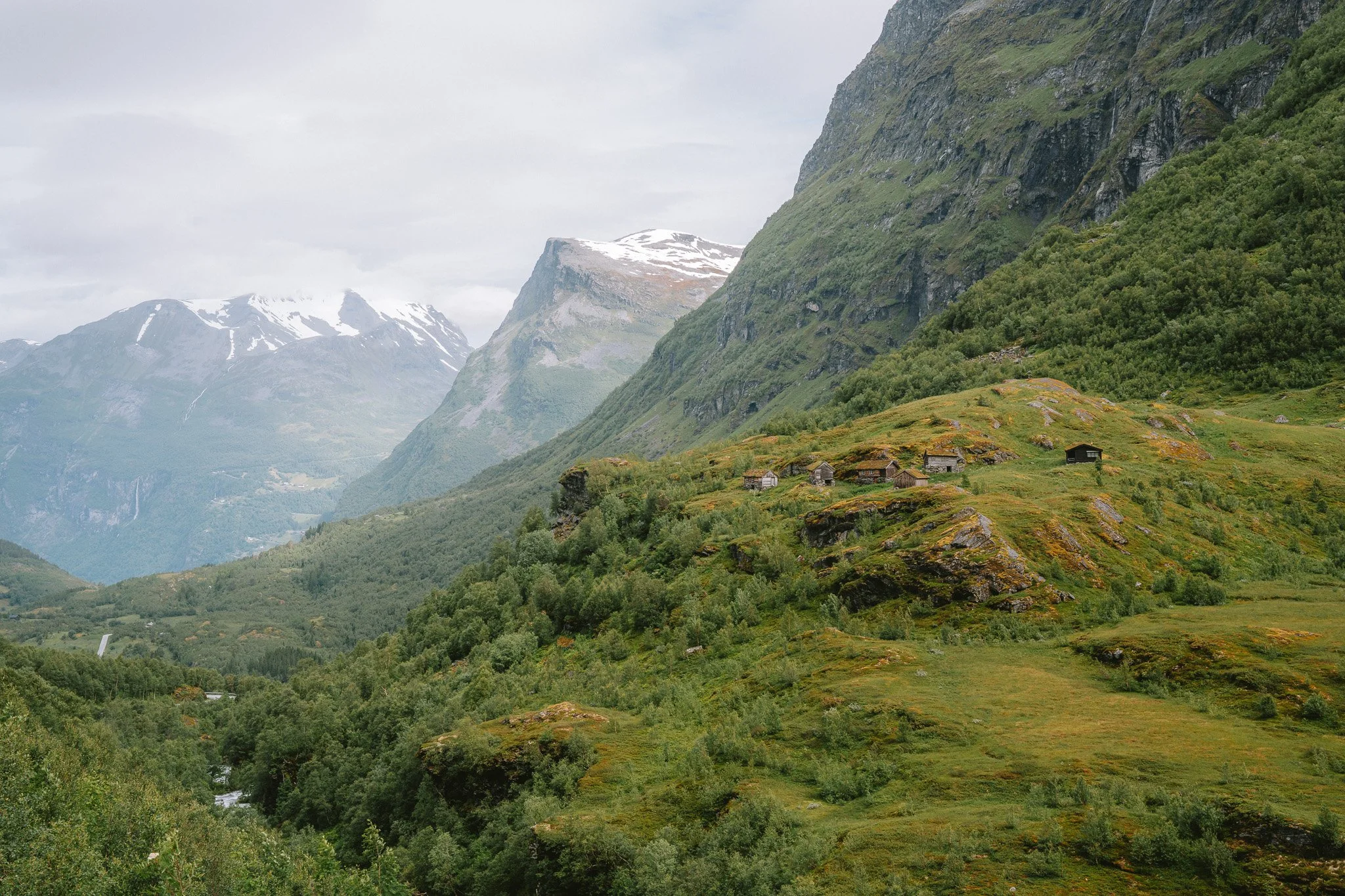 Mountain landscape with lush green hills, small wooden houses scattered on the hillside, and snow-capped peaks in the background under cloudy sky.