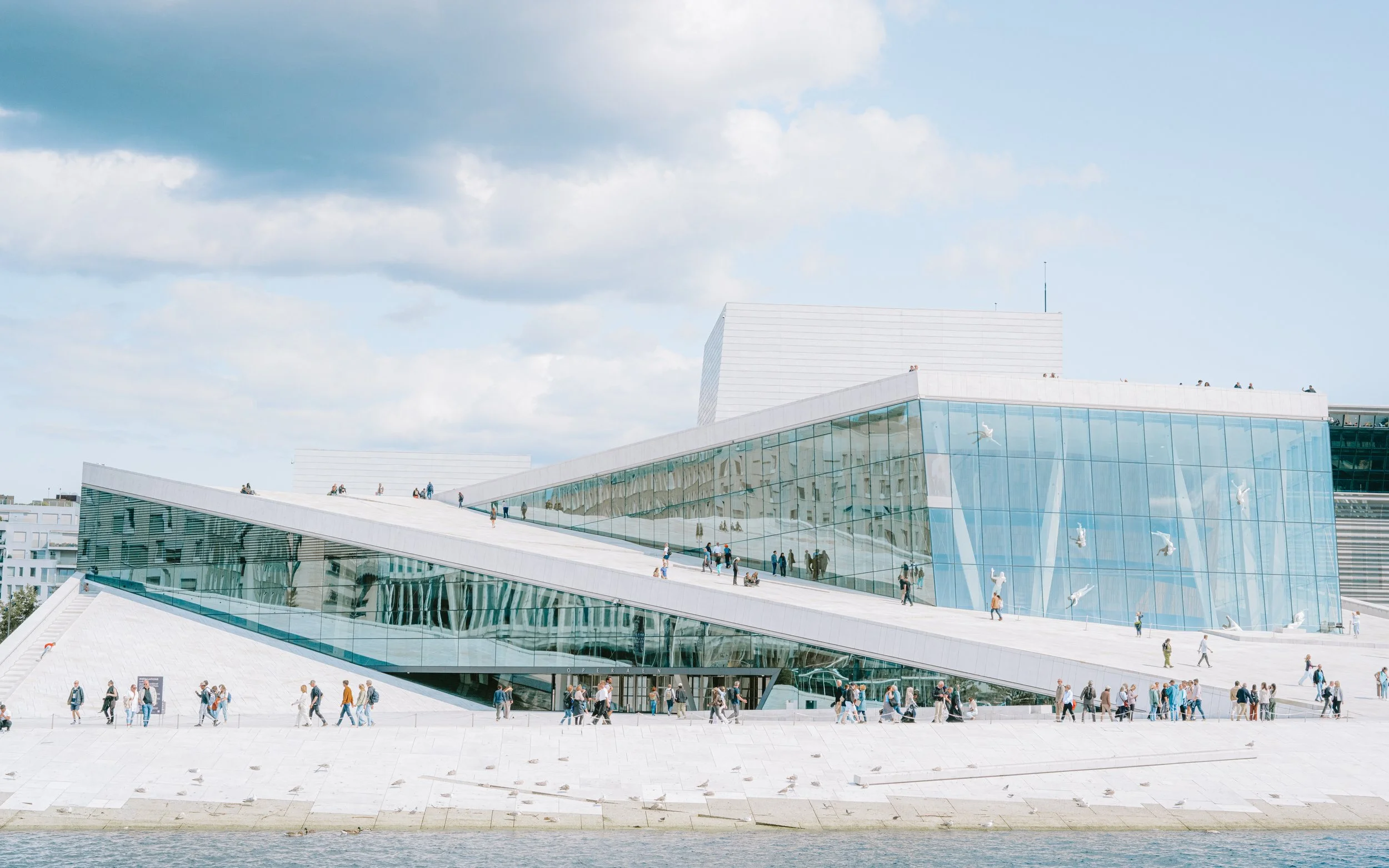 Modern building with glass facade and sloped white roof, with people walking on the roof and outside, near a body of water, under a partly cloudy sky.