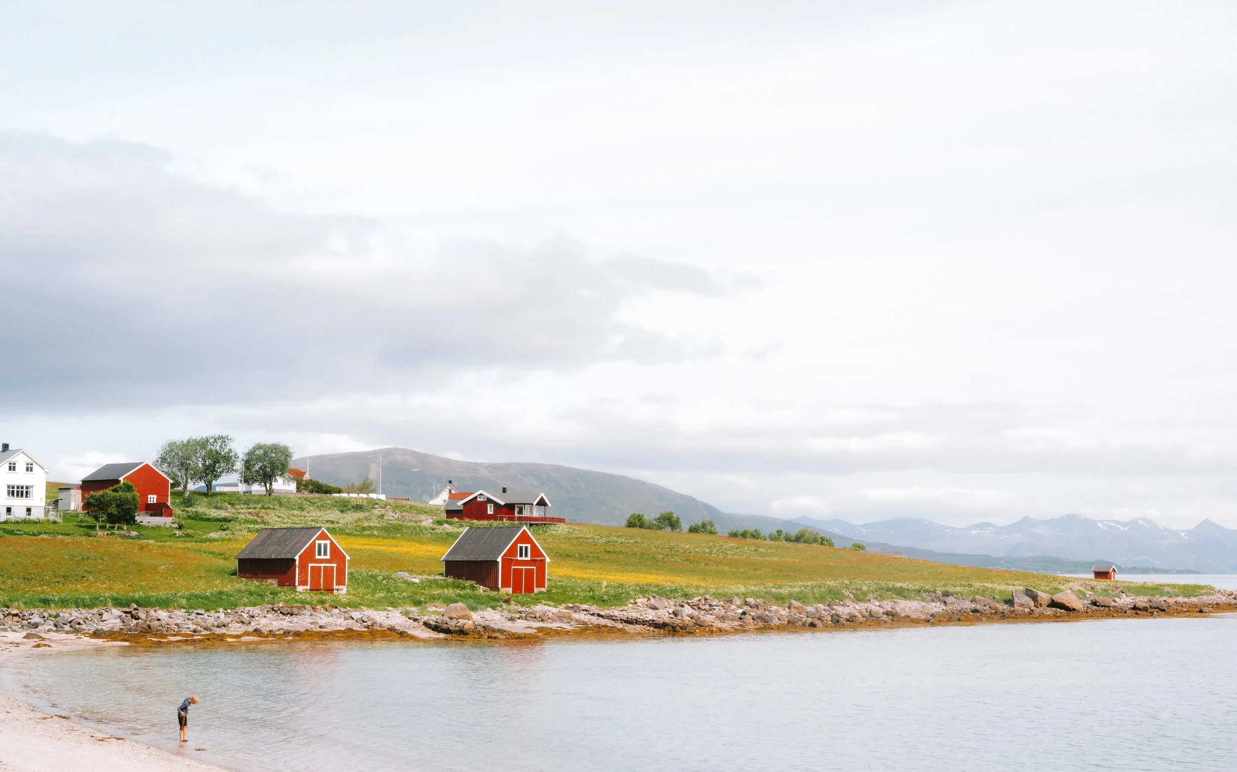 Coastal landscape with red and white houses and small boats, mountains in the background, and a person wading in the water.