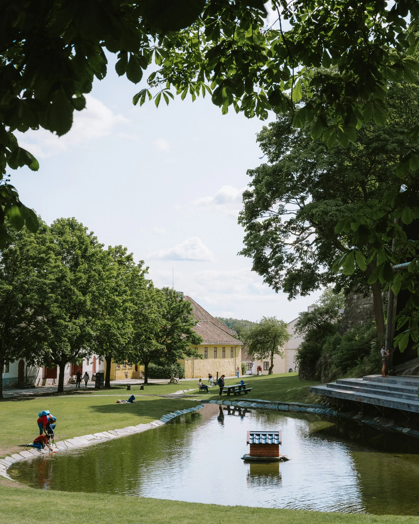 A park scene with a pond, trees, benches, and people relaxing and walking, under a partly cloudy sky.