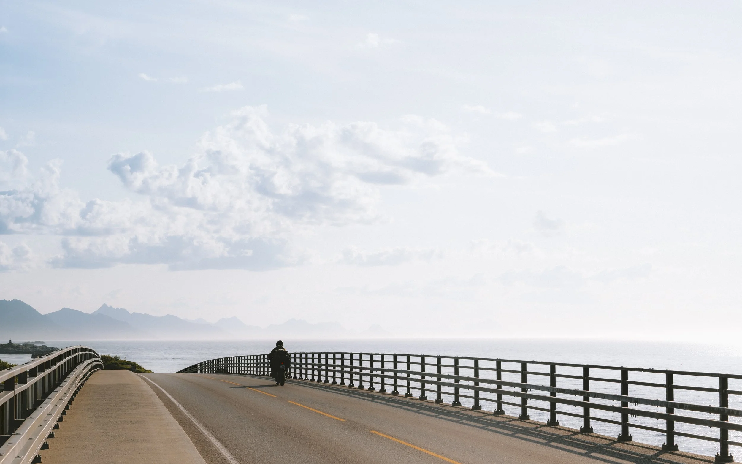 A person riding a motorcycle on a coastal road with ocean on the right and mountains in the distance on the left.