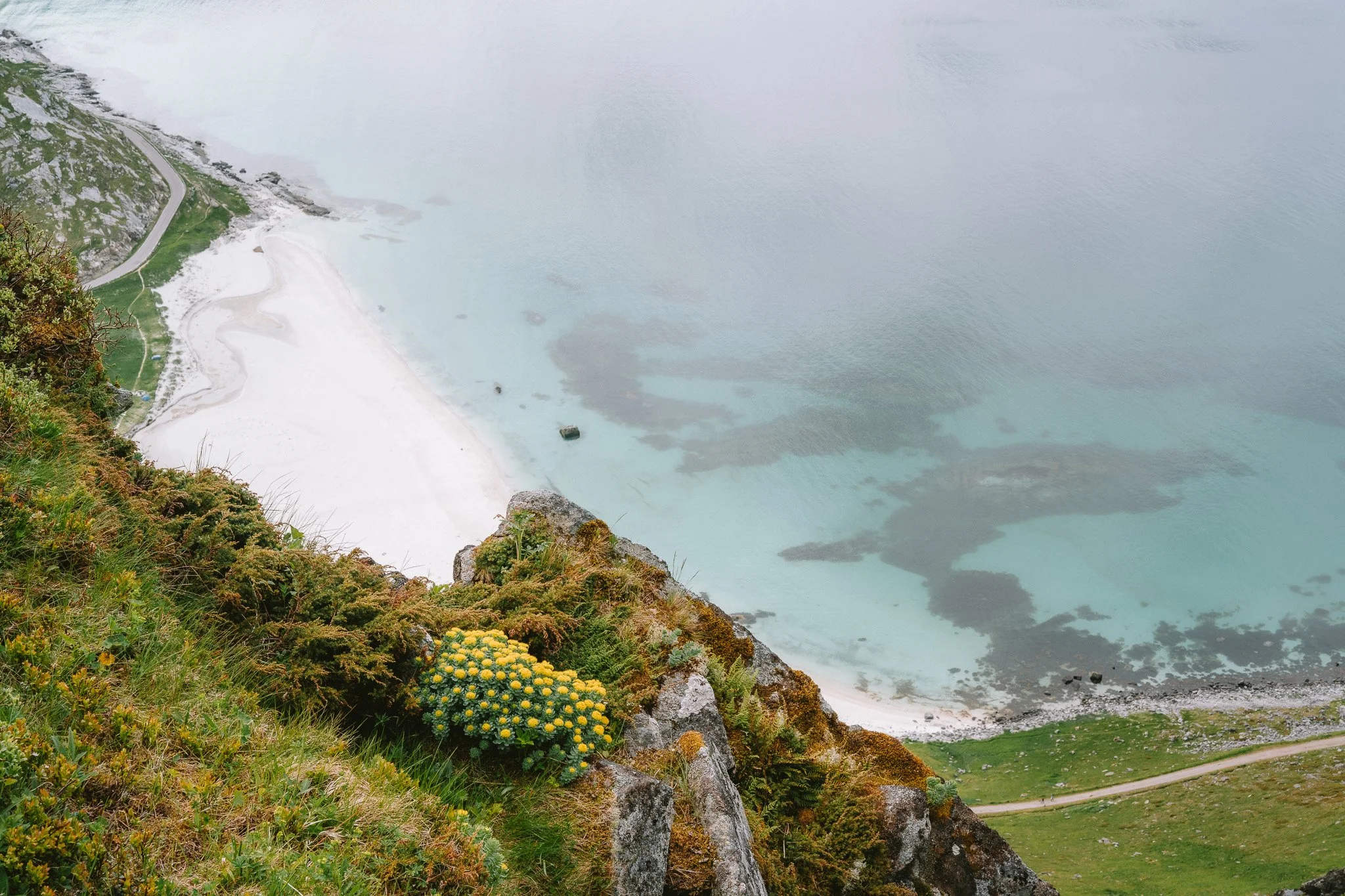 Aerial view of a coastal landscape showing a sandy beach, turquoise water, rocky shoreline, green vegetation, and a winding road.