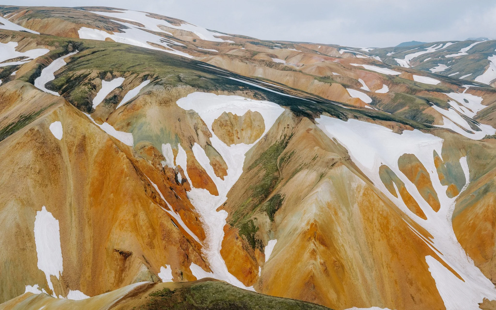 Colorful mountain landscape with patches of snow on rugged, multicolored peaks under a cloudy sky.
