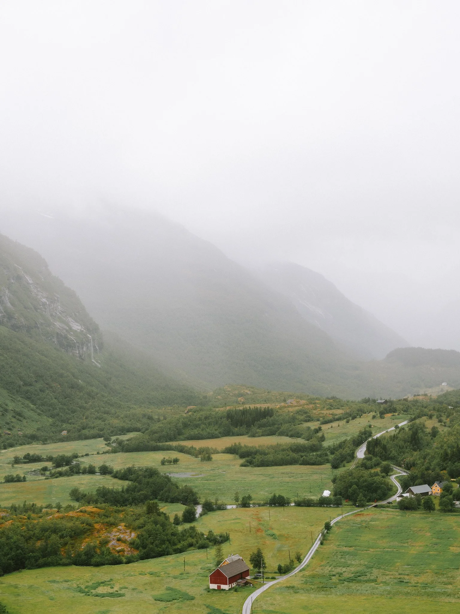 Scenic view of a foggy green valley with rolling hills, a winding road, a red barn, and scattered houses.