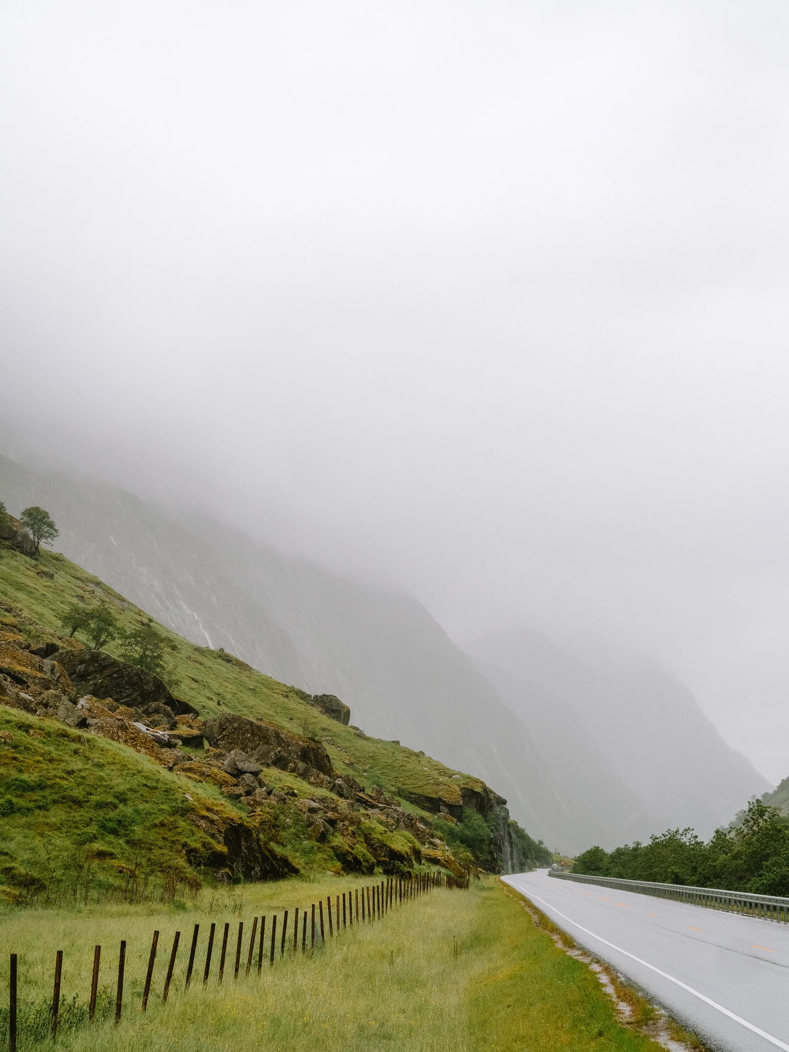 A winding highway along a lush, green hillside with a wooden fence on the left and fog-covered mountains in the background.