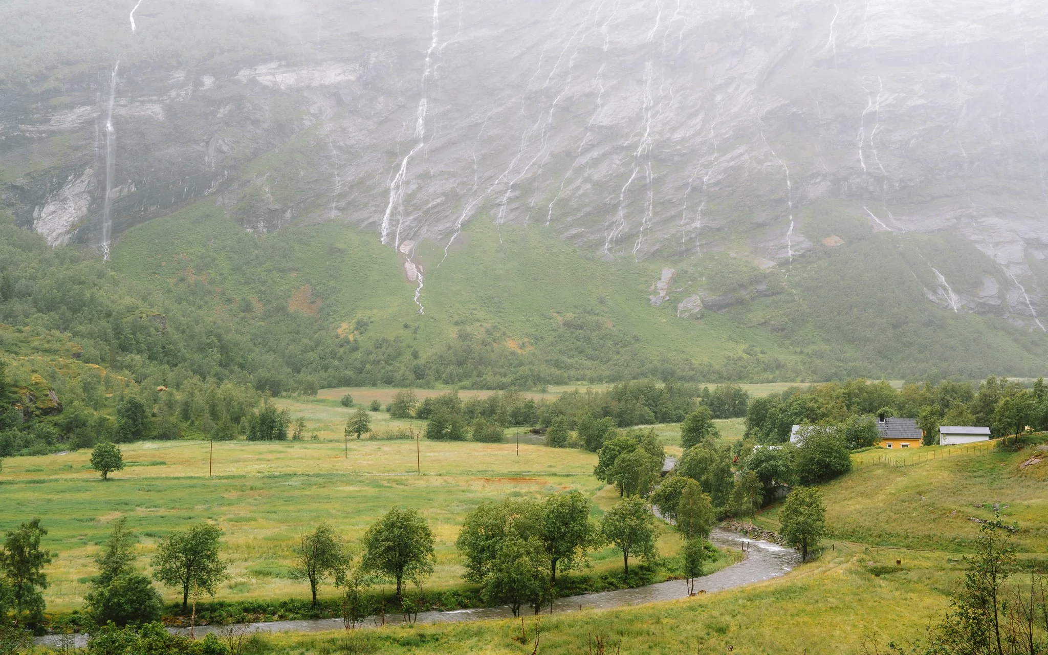 A scenic landscape with a large mountain in the background, featuring rain streams and waterfalls, lush green trees, a river flowing through the grassy fields, and a few houses with gray and yellow exteriors.