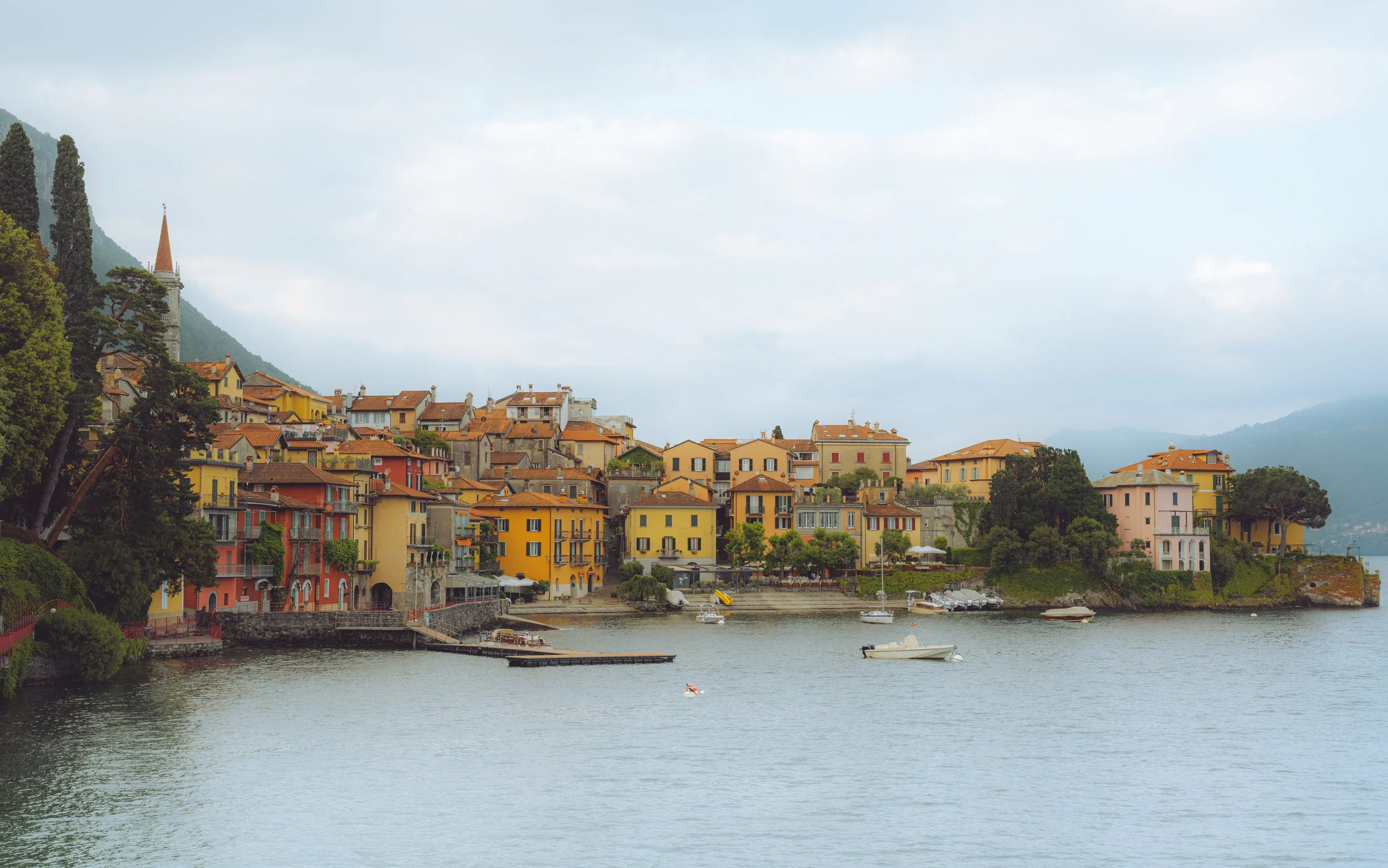 Colorful houses along a lakeshore with boats in the water and mountains in the background.