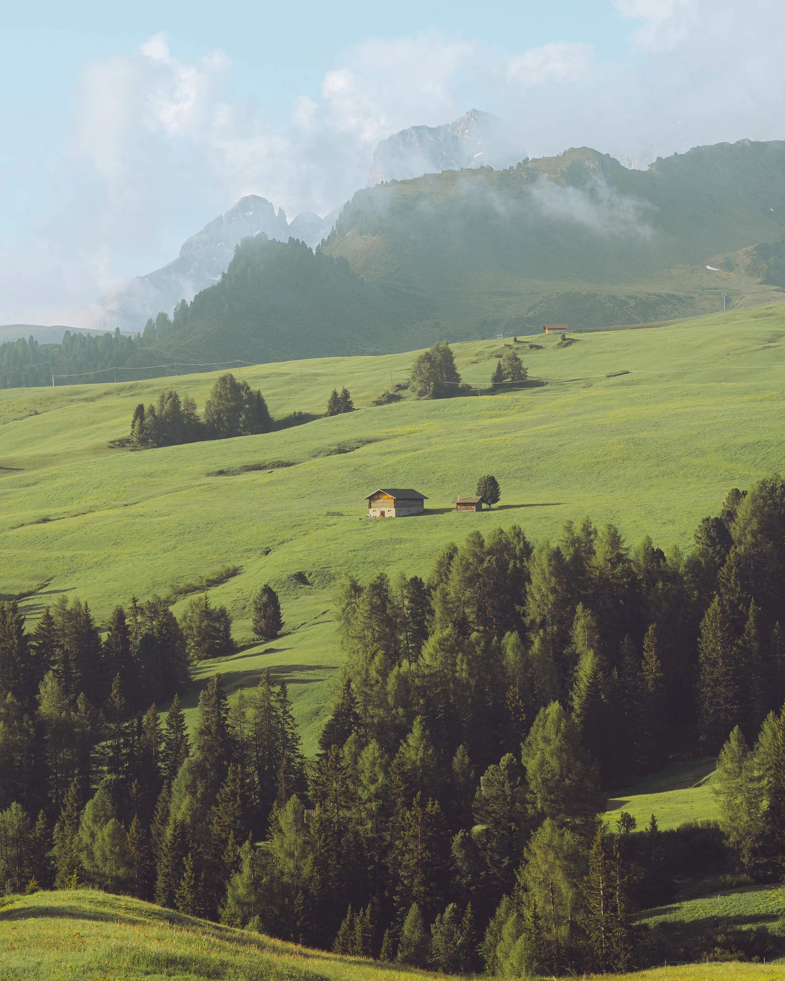 Green rolling hills with scattered trees and small houses, mountain peaks in the background under a partly cloudy sky.